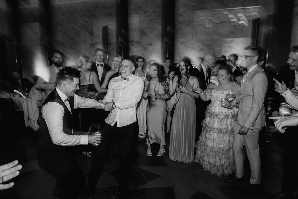 Groom dances with guest as wedding crowd cheers in black and white photo.