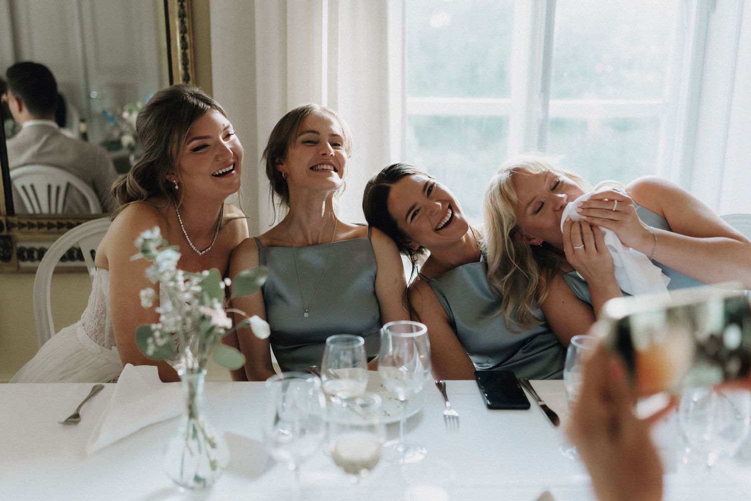Four bridesmaids laughing joyfully together at a wedding reception table in a bright elegant venue.