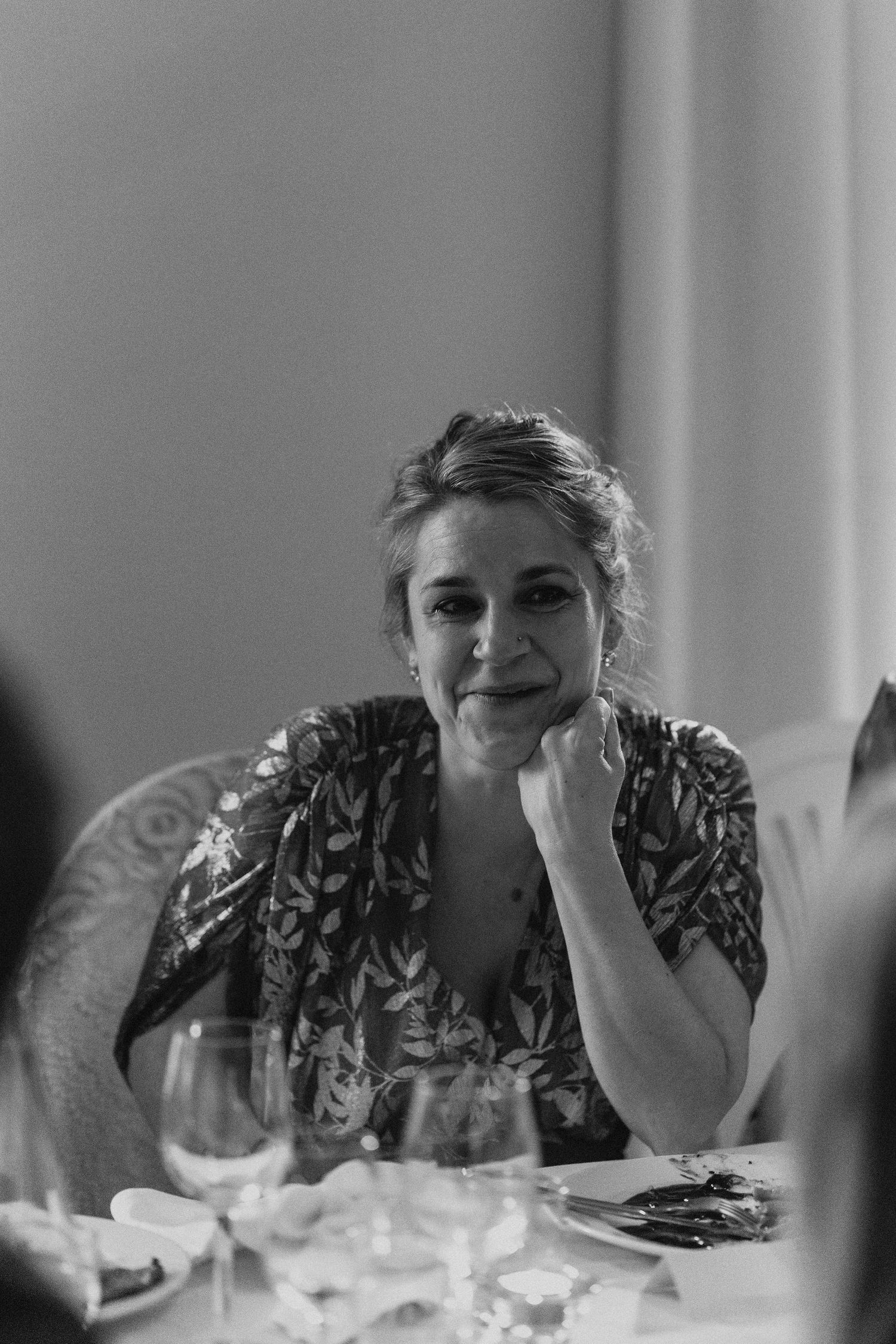 Elegant woman in floral dress resting chin on hand at wedding reception dinner table, black and white photo.