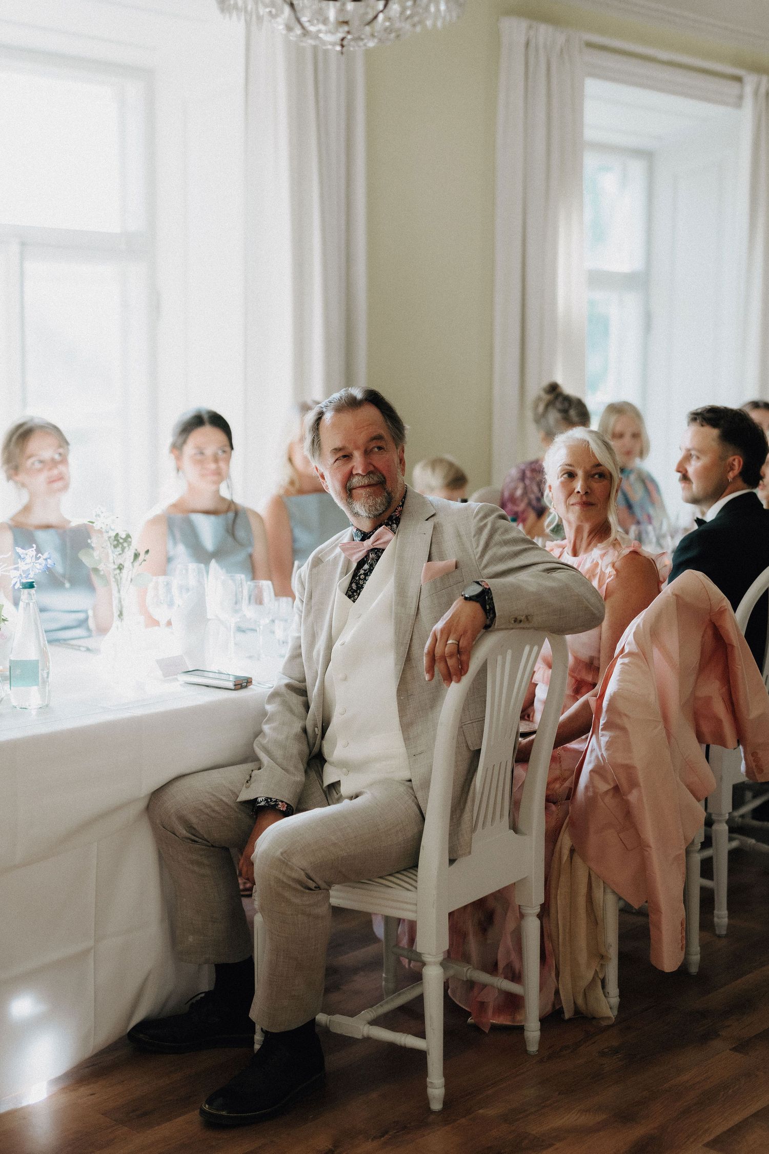Smiling wedding guest in beige suit sits relaxed at reception table surrounded by other guests.