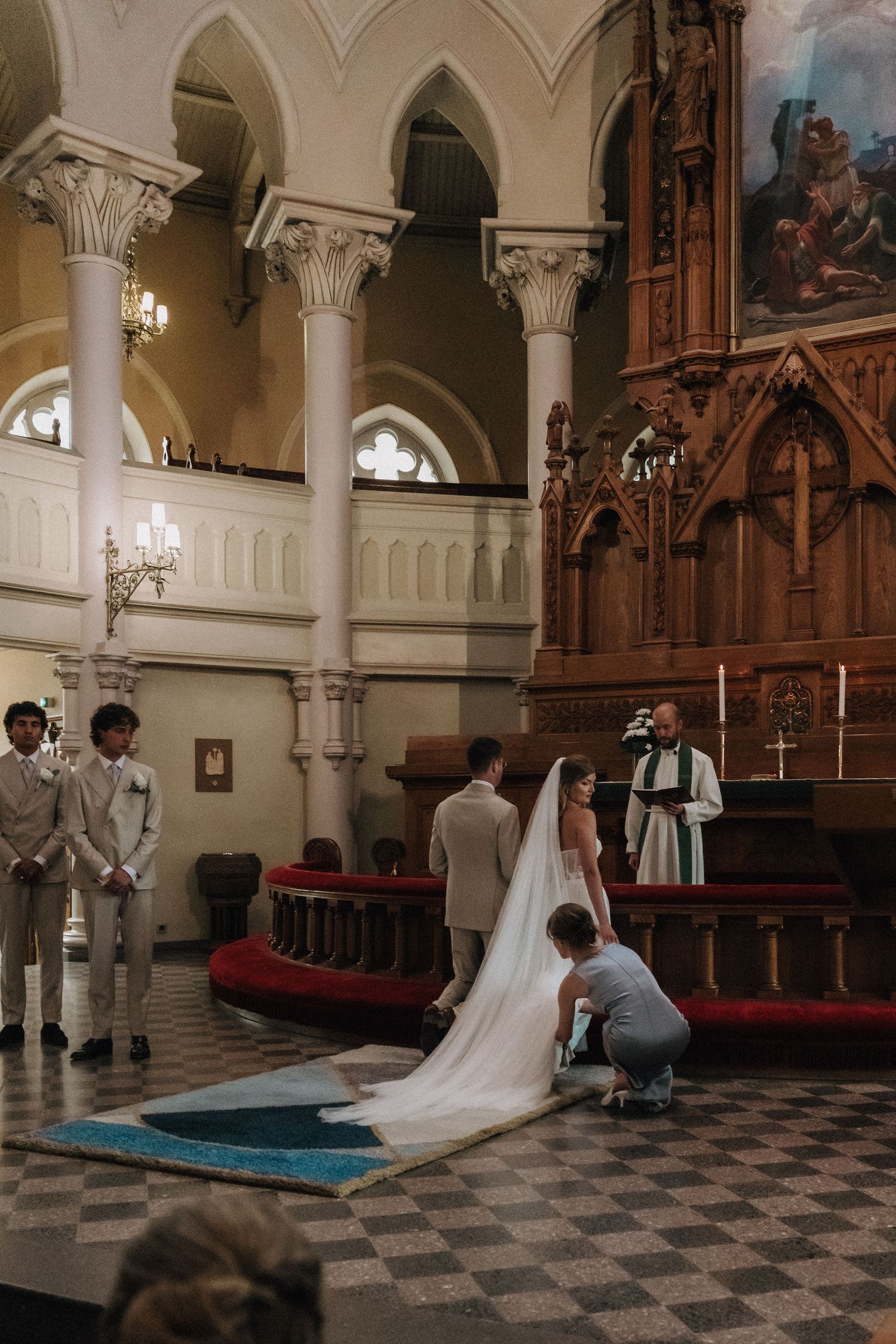 Bridesmaid adjusts bride's cathedral veil at church altar while officiant and groom wait.