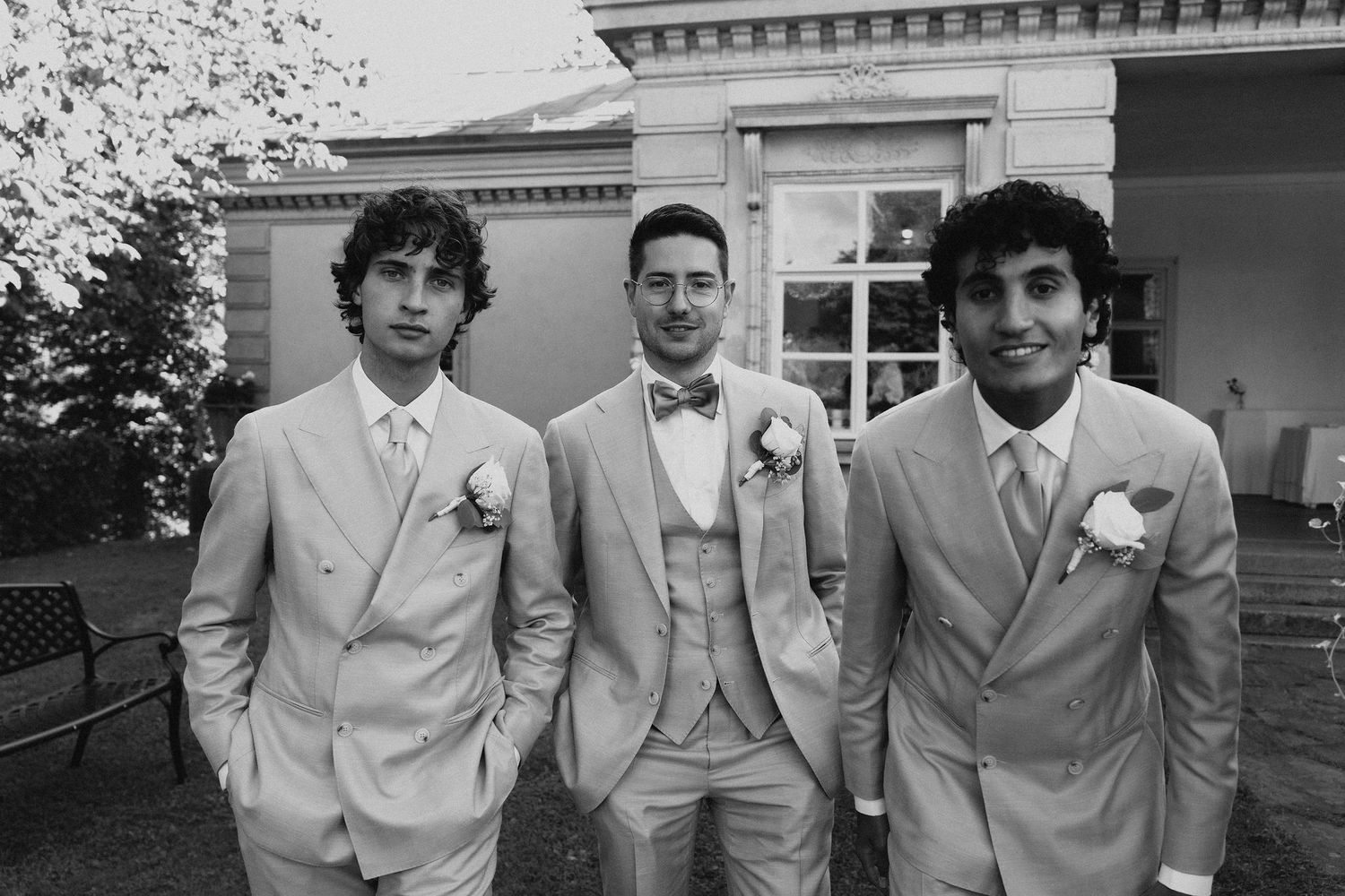 Black and white photo of three groomsmen in light suits with boutonnieres at outdoor wedding.