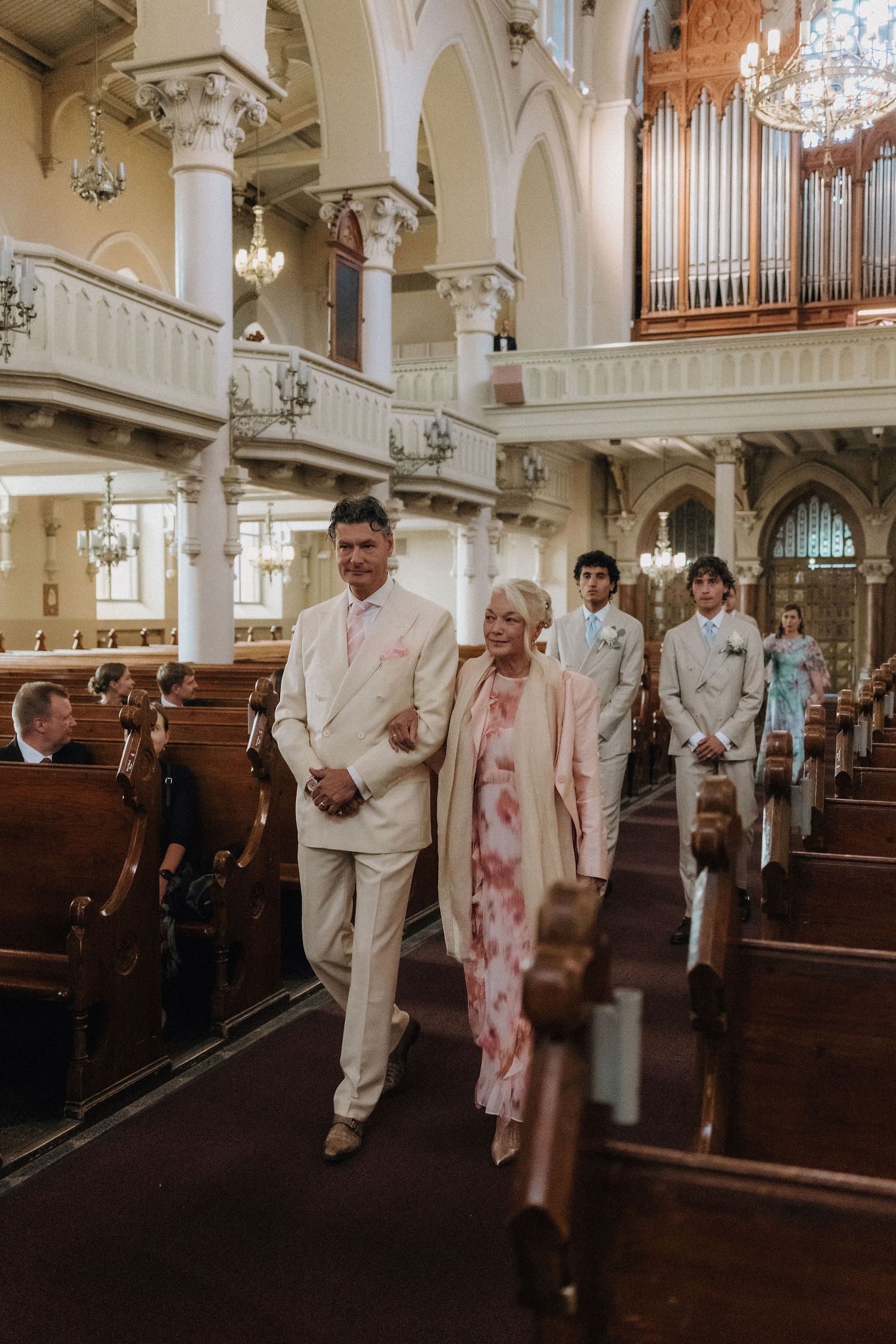 Groom escorts mother down church aisle in elegant wedding processional with pipe organ backdrop.