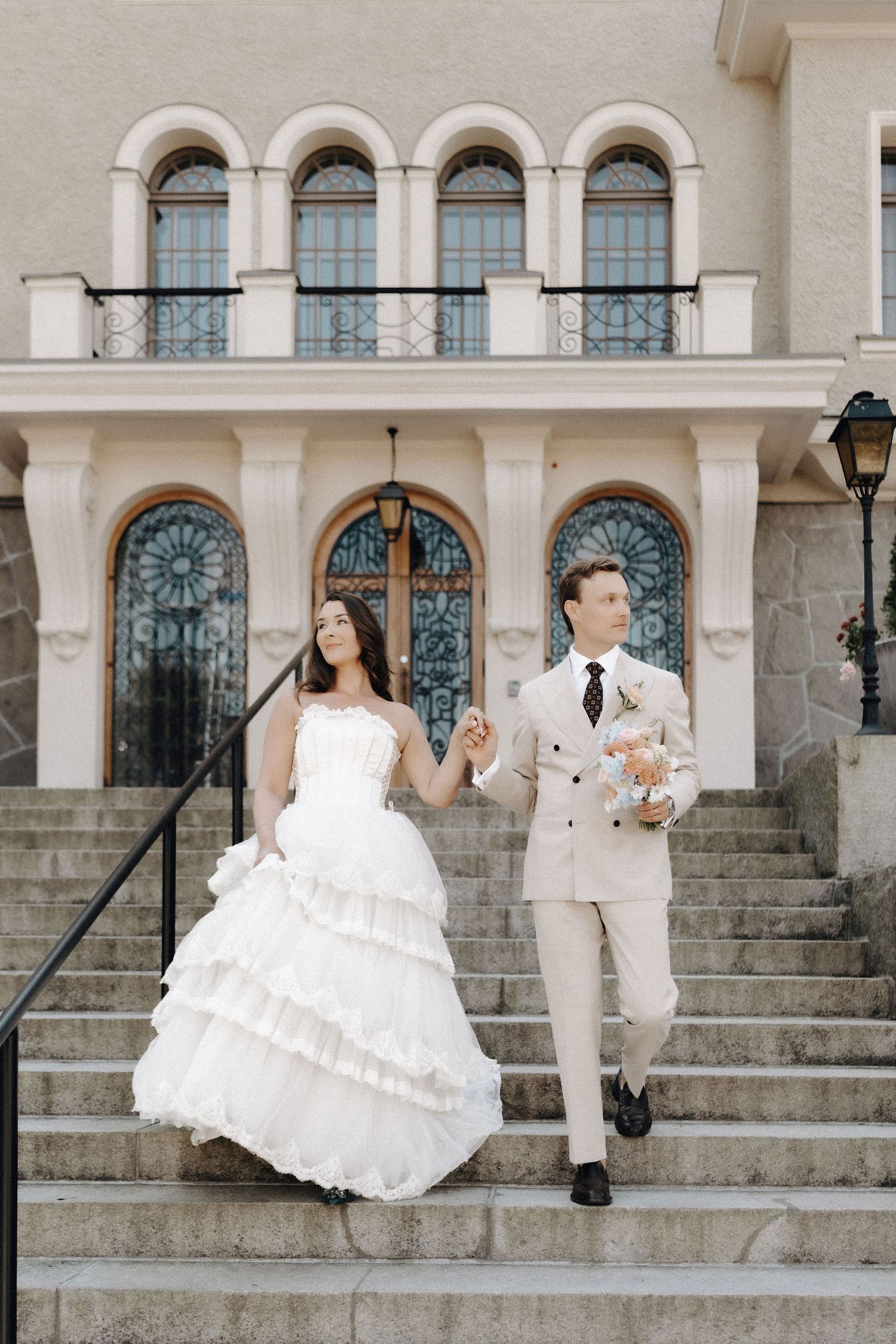 Bride and groom walking down elegant stone steps outside a grand building with arched stained glass windows.