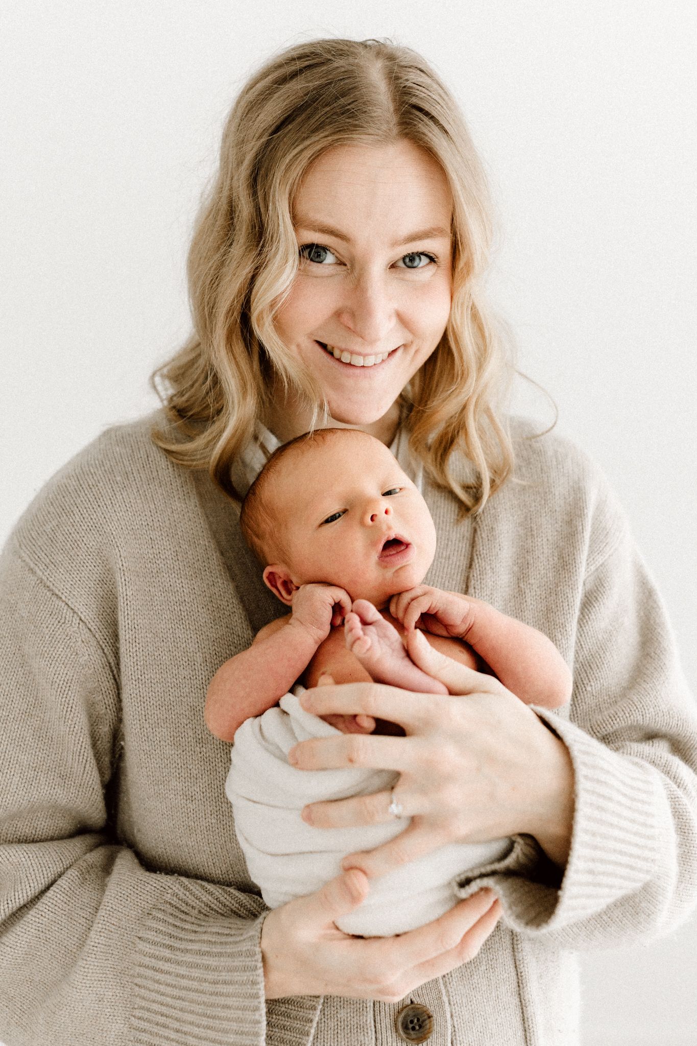 Smiling blonde mother in beige cardigan holding her alert newborn baby against a white background.