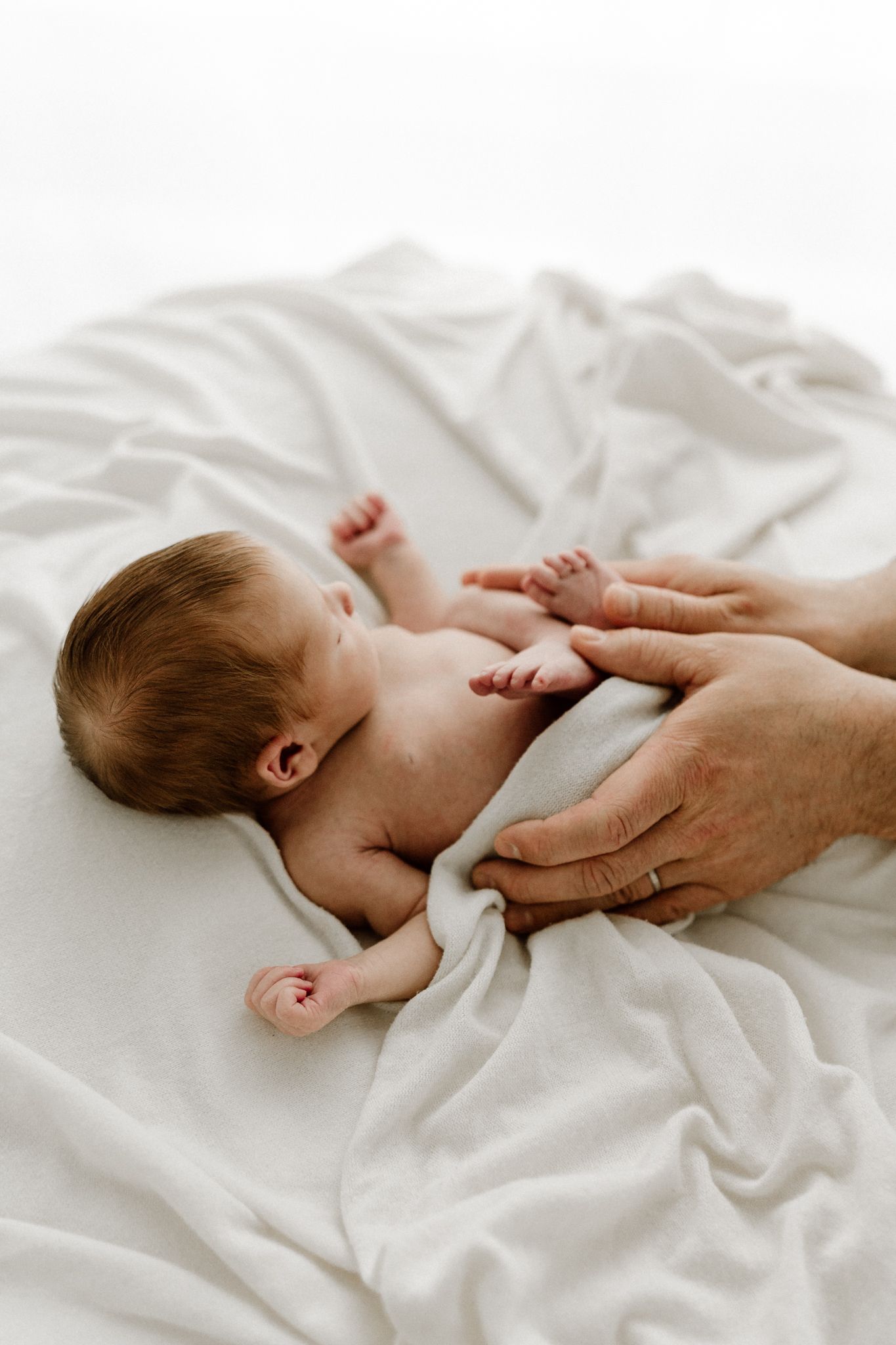Parent gently holds sleeping newborn baby lying on soft white blanket during an intimate newborn photo session.