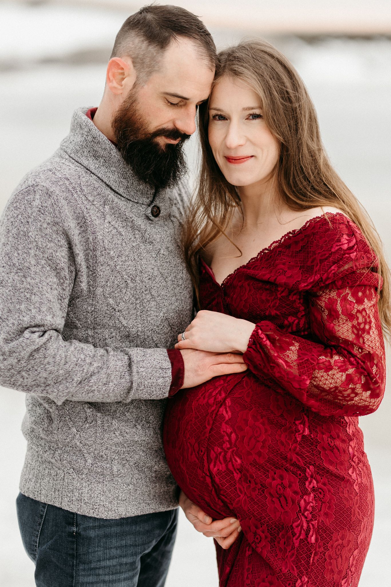Couple in maternity photo shoot outdoors in winter, woman in red lace dress cradling baby bump.