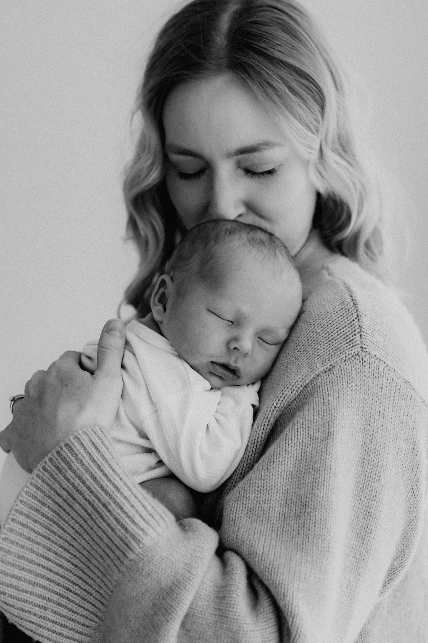 Black and white photo of a mother tenderly kissing her sleeping newborn baby while cradling it close.