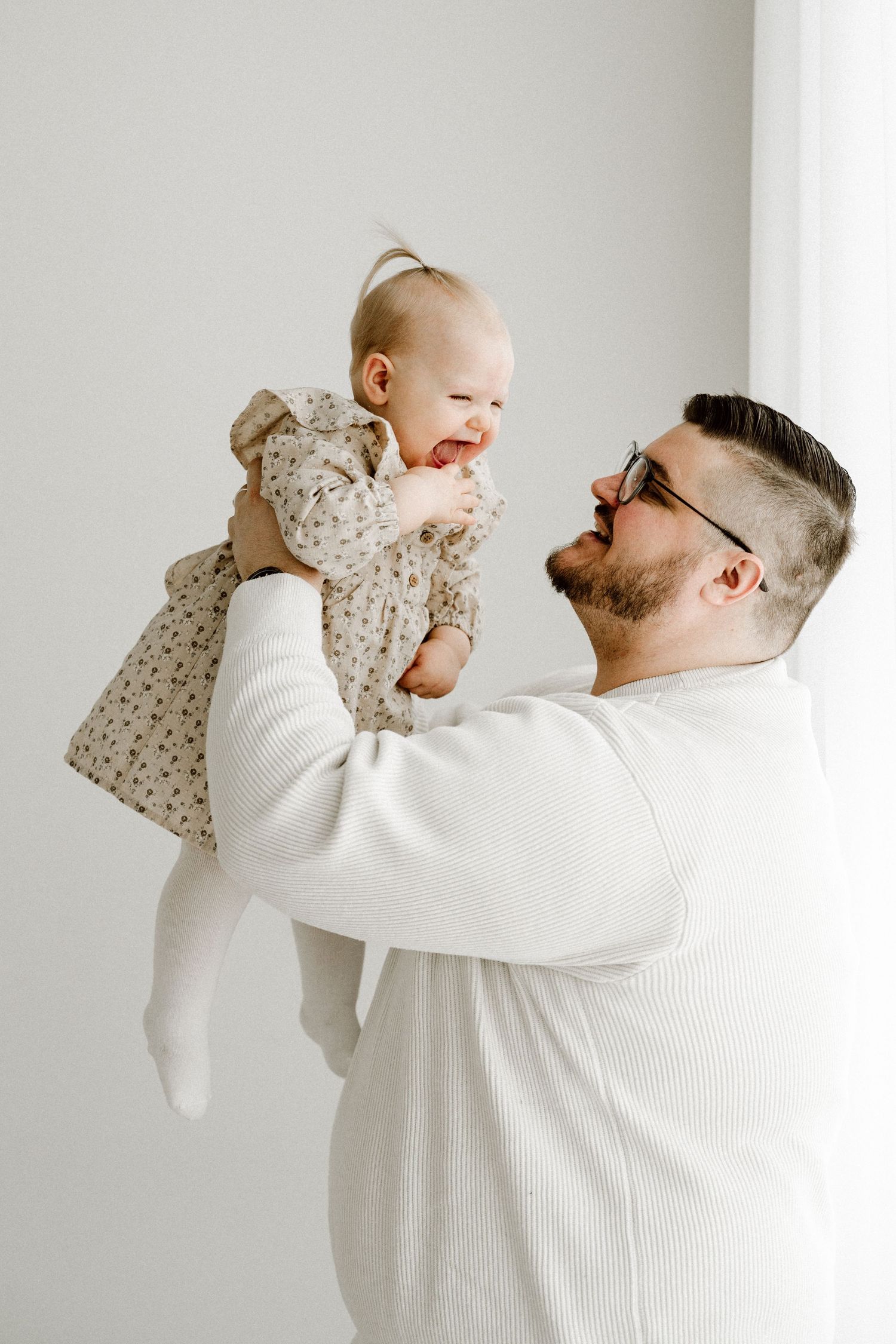A bearded father in glasses lifts his laughing baby girl in a floral dress in a bright white studio.