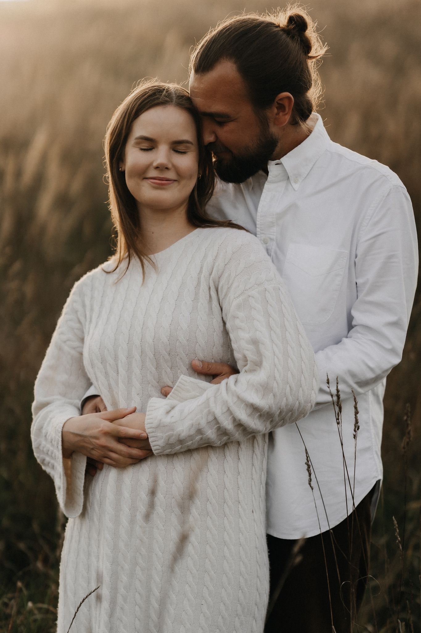 Man with hair bun embracing woman in white knit sweater from behind in a golden sunlit field.