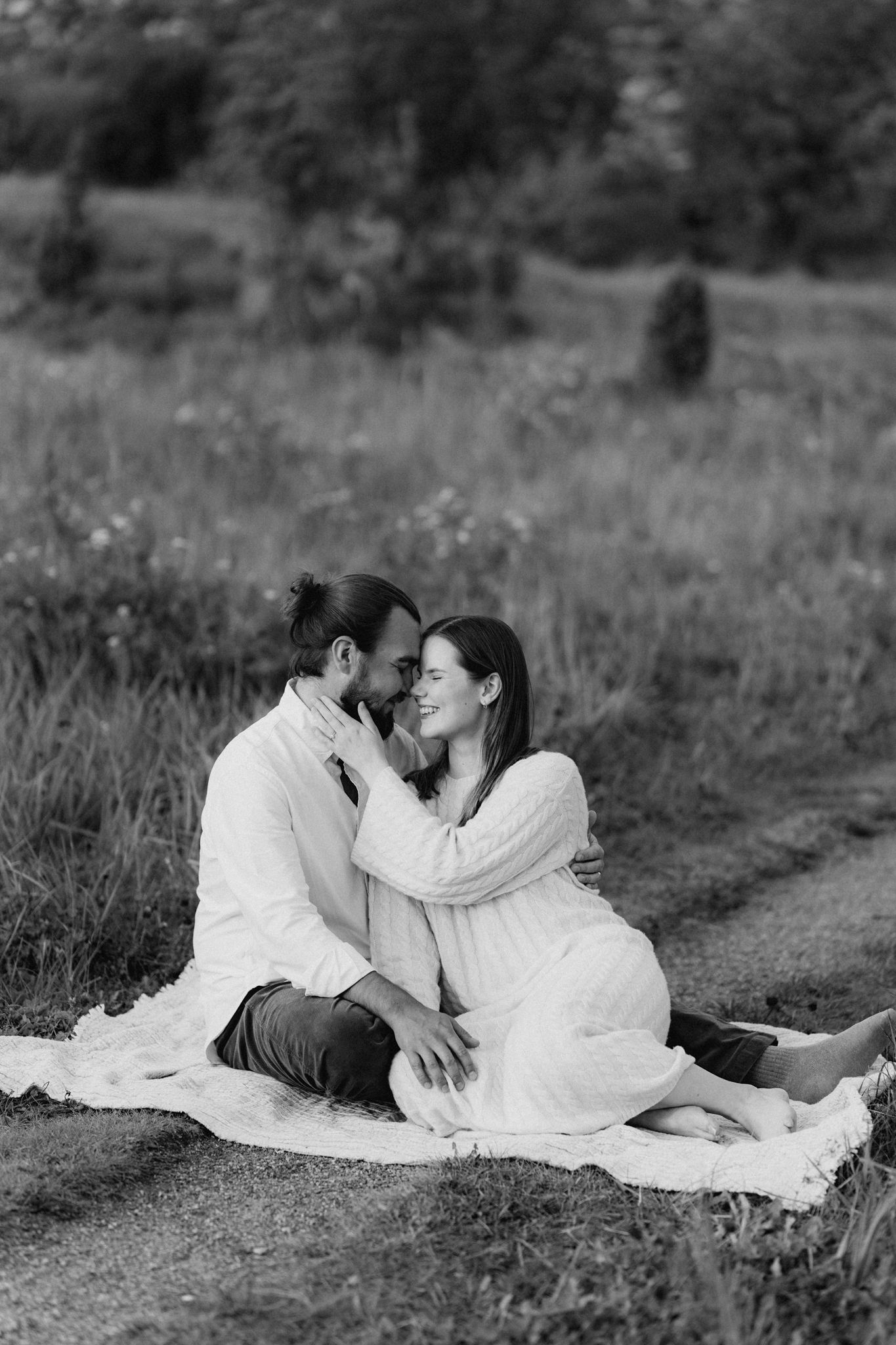 Black and white photo of a couple sitting on a blanket in a field, sharing a tender moment nose-to-nose.