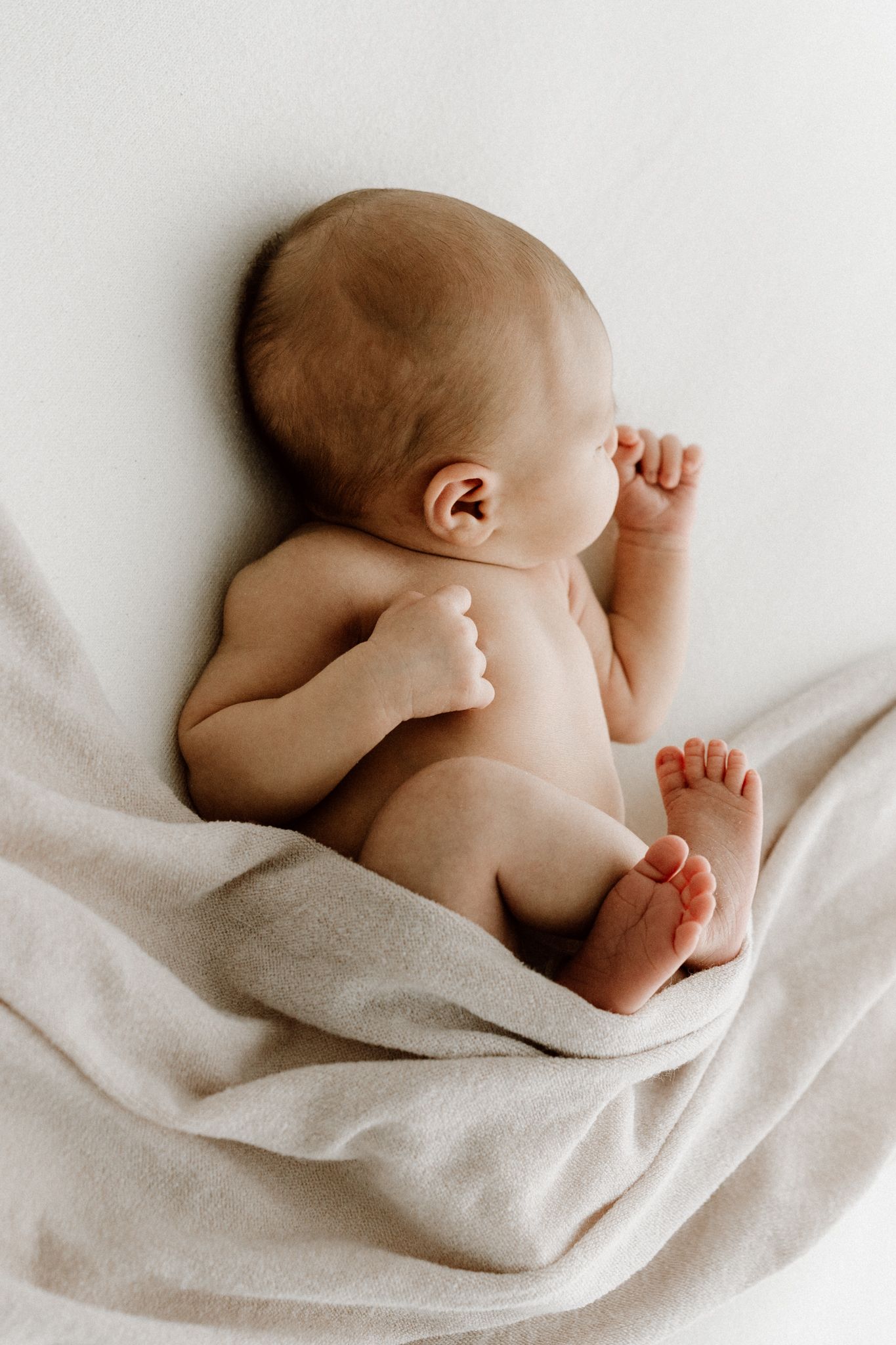Newborn baby lying on soft beige blanket, viewed from behind showing tiny feet and hands.
