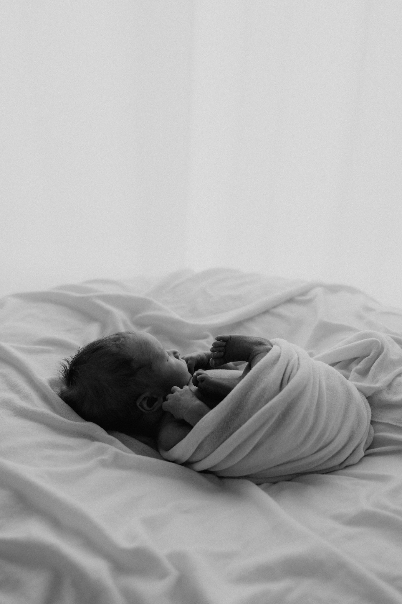 Black and white photo of a swaddled newborn baby resting on white bed sheets.