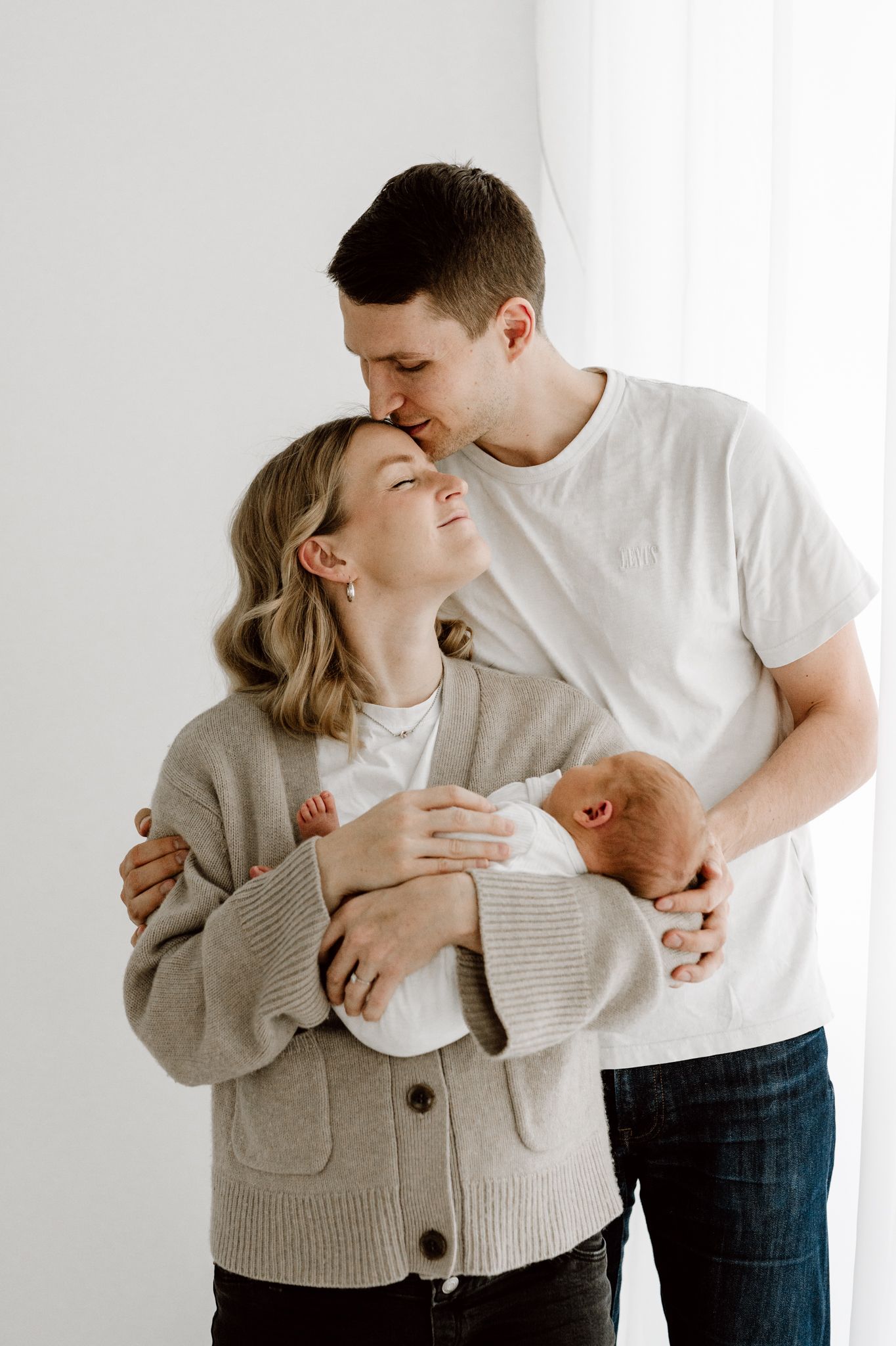A father kisses the mother on the head as she holds their newborn baby in a bright studio setting.