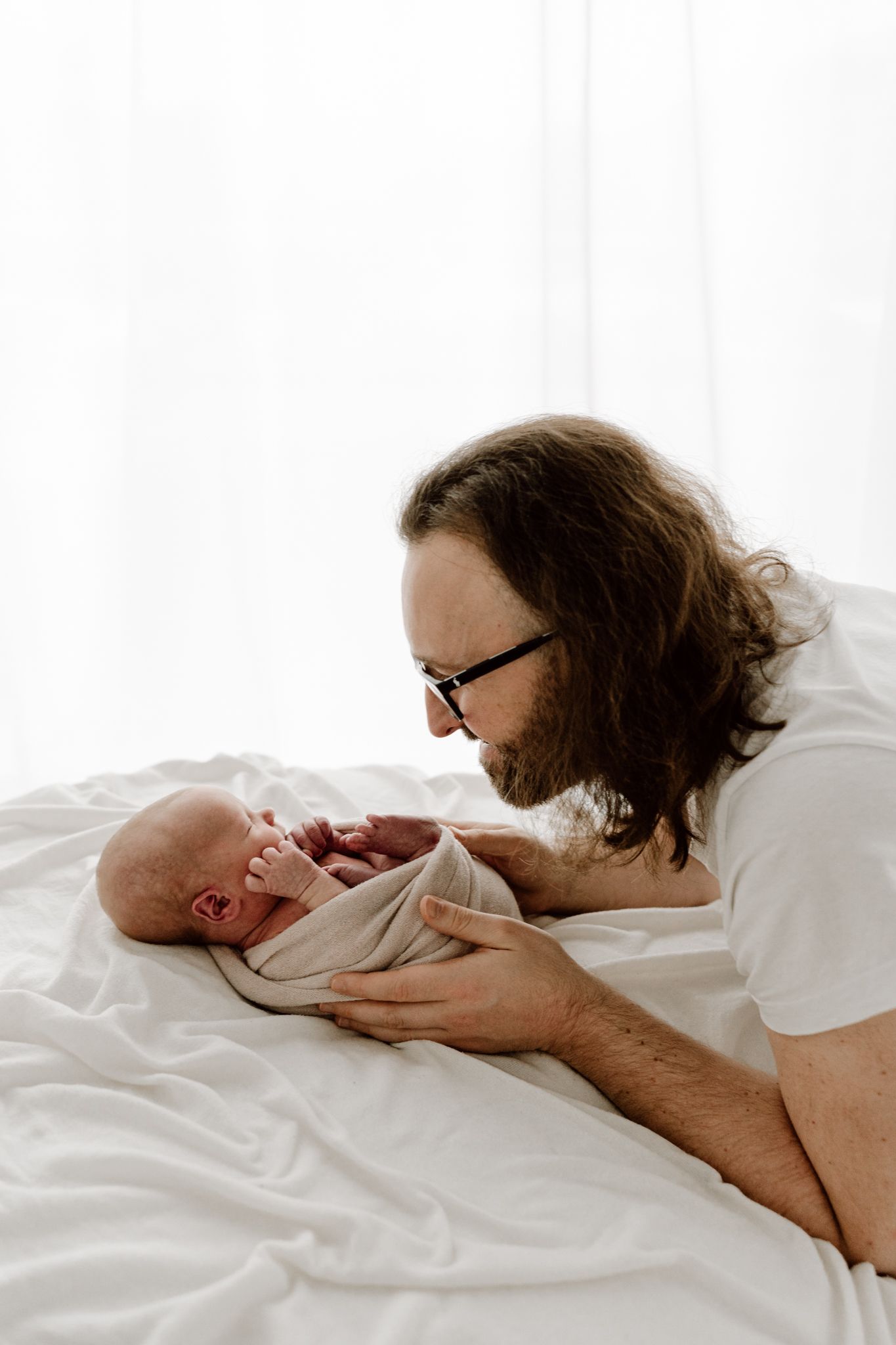 Father with glasses lying on white bedding, smiling at his crying swaddled newborn baby.