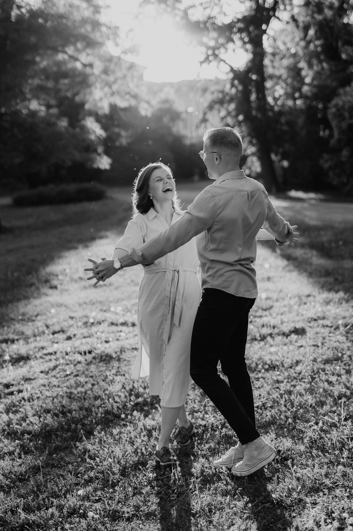 Black and white photo of a laughing couple dancing playfully on a grassy park lawn.