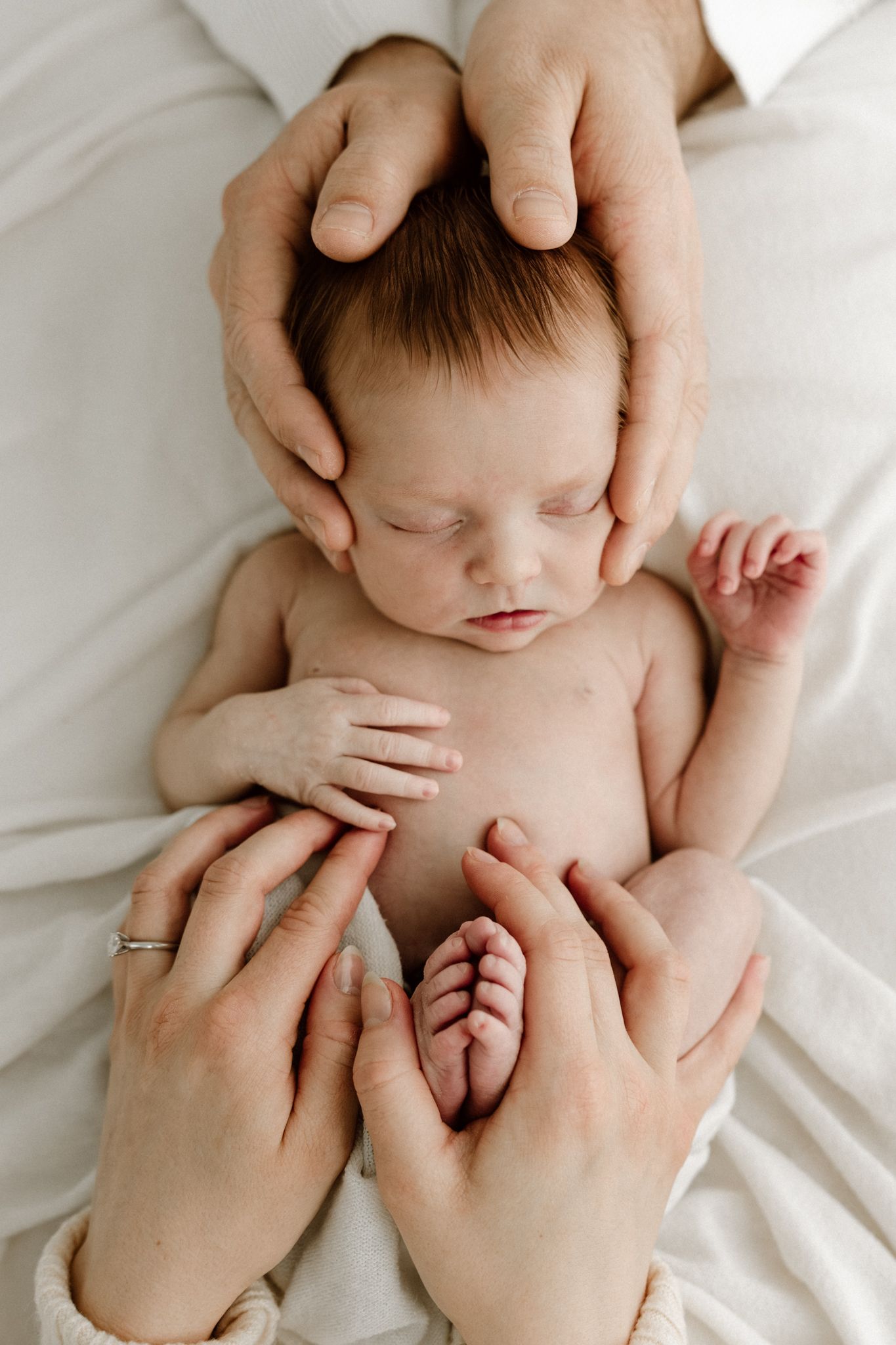 Parents tenderly cradle sleeping newborn baby with gentle hands on white blanket.