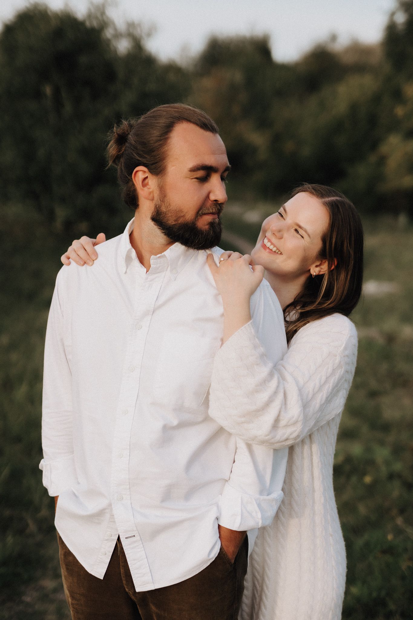 A smiling woman in white leans affectionately on her bearded partner outdoors.