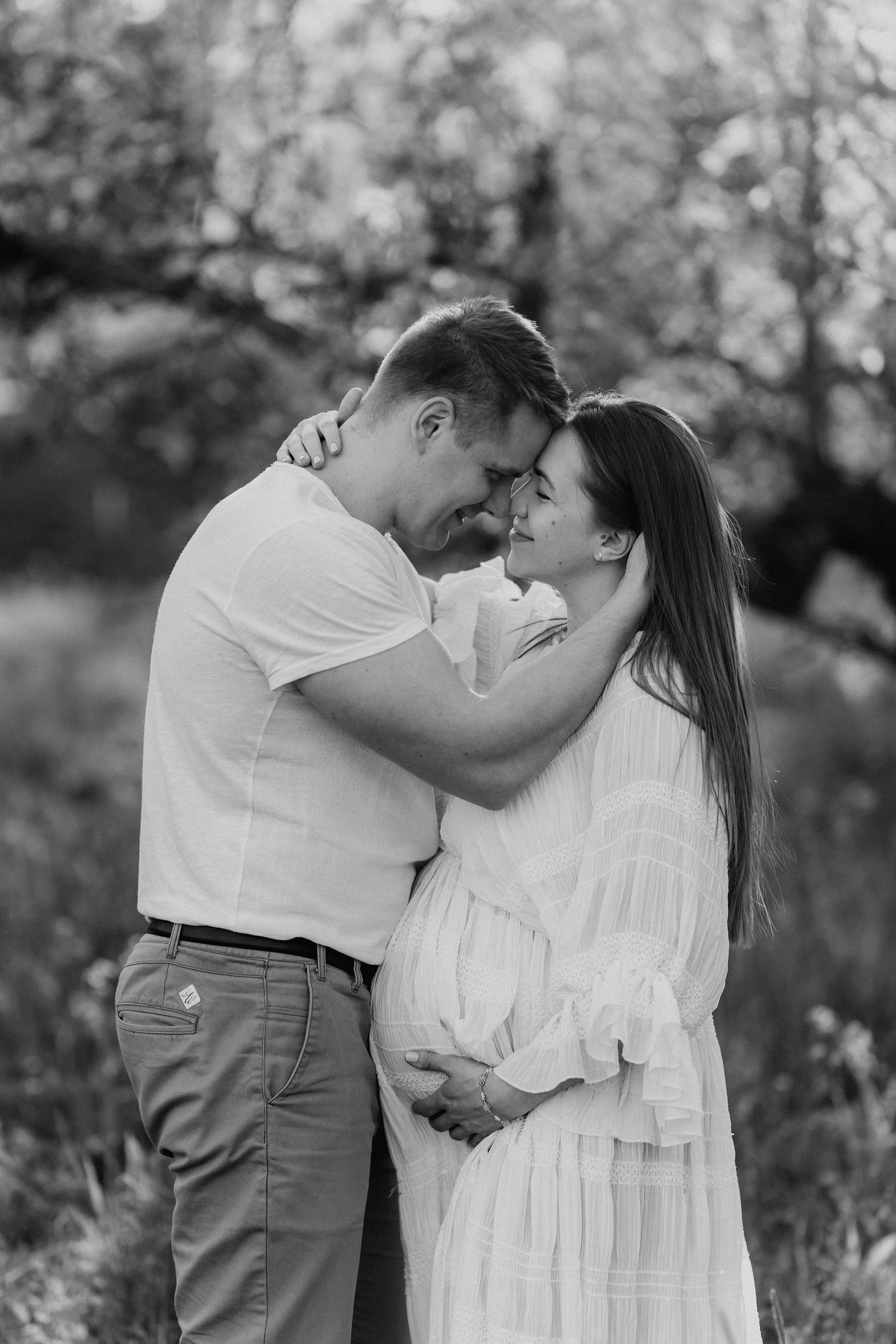 Black and white photo of couple embracing tenderly, pregnant woman in white dress outdoors.