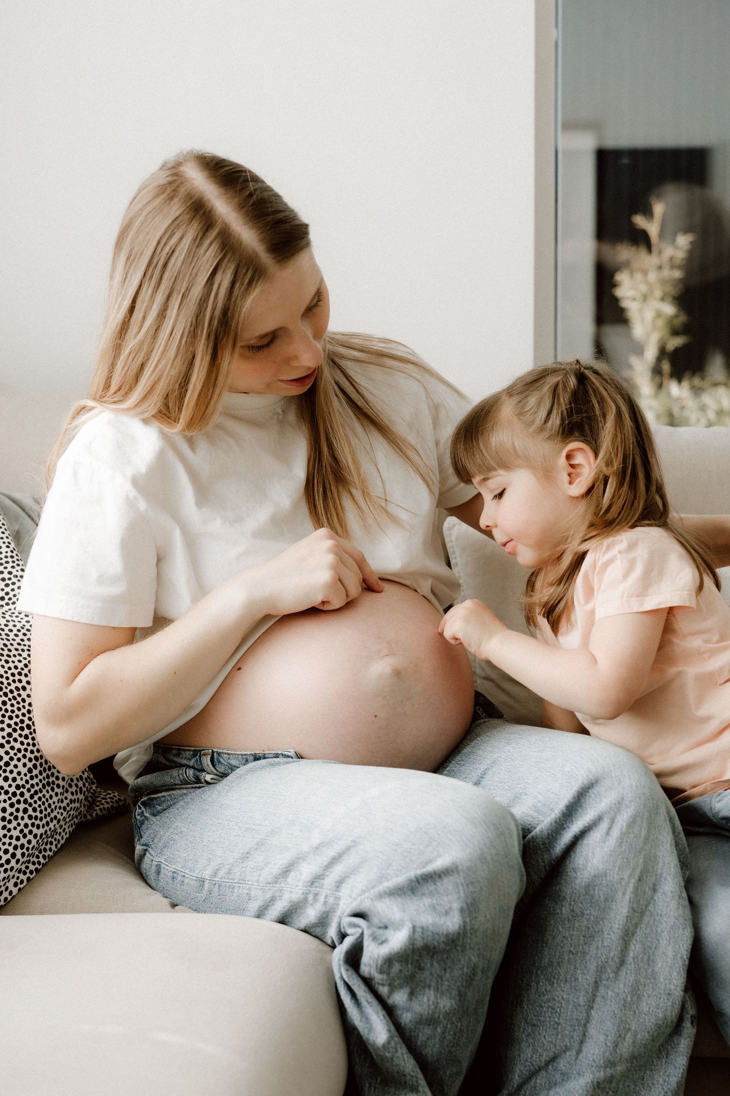 Pregnant woman in white shirt and jeans sitting with young daughter touching bare baby bump.