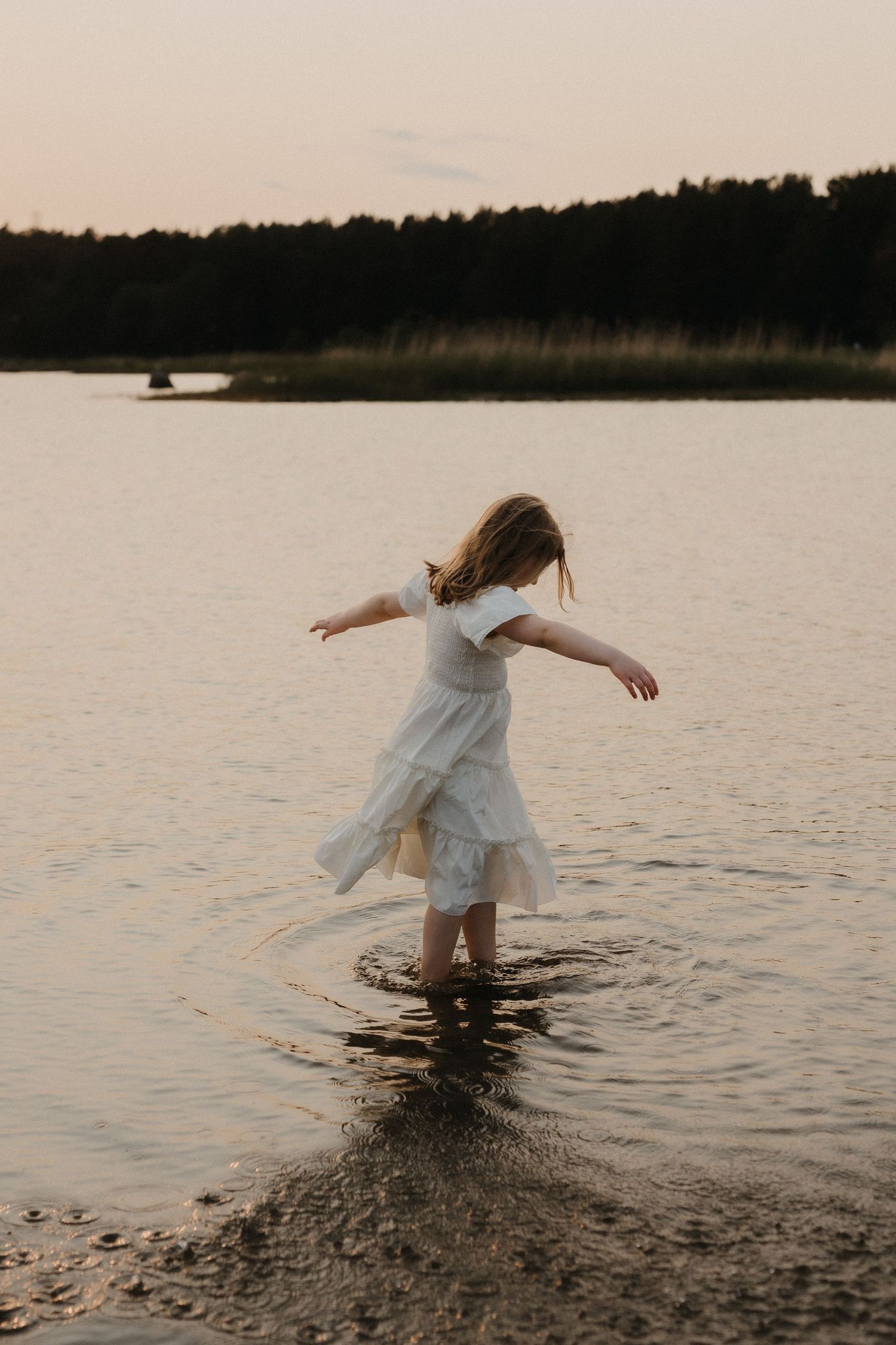 A young girl in a white dress twirls gracefully in shallow lake water at dusk.