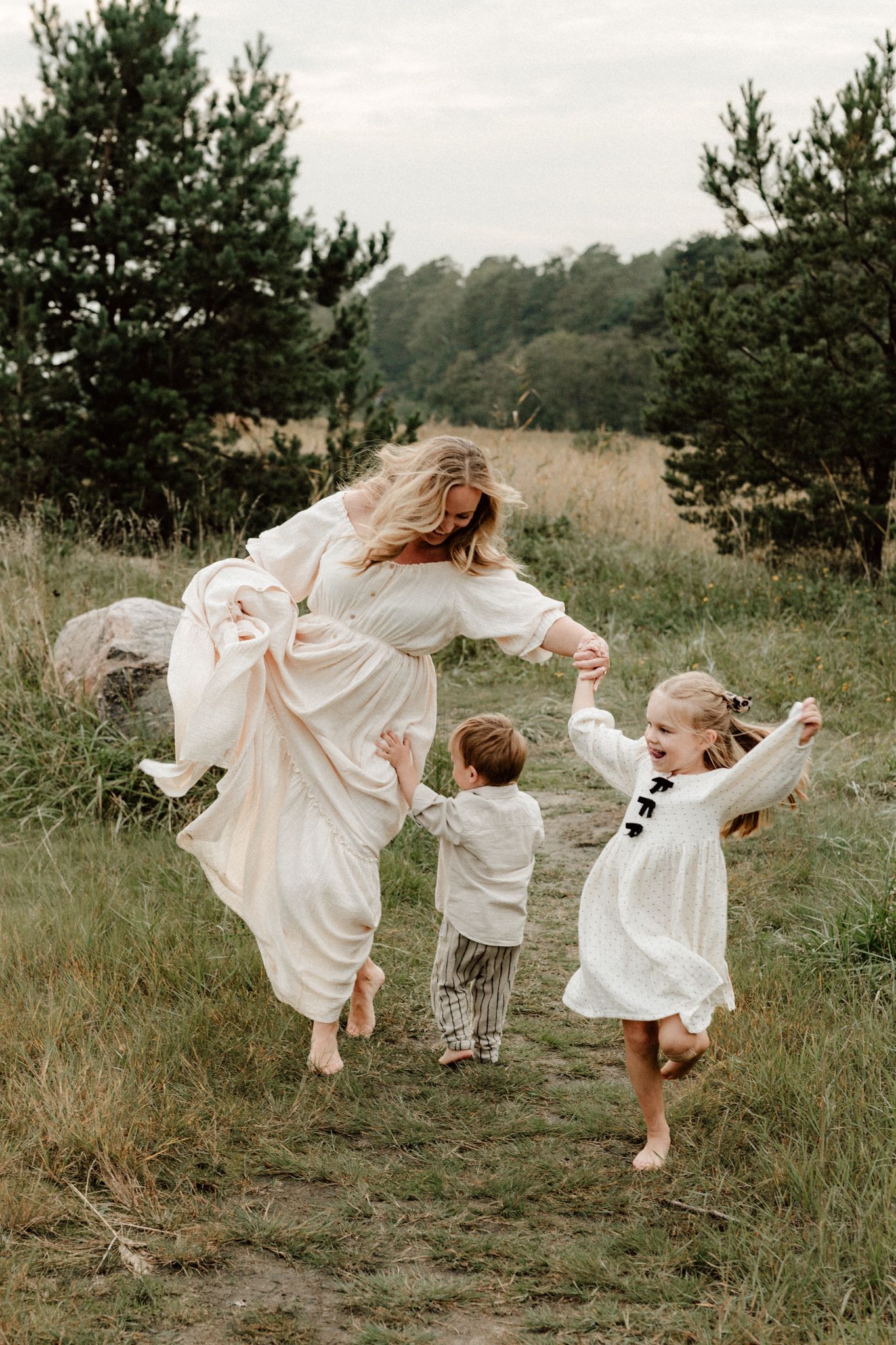 A joyful mother in a flowing cream dress dances with her two young children in a lush green meadow.
