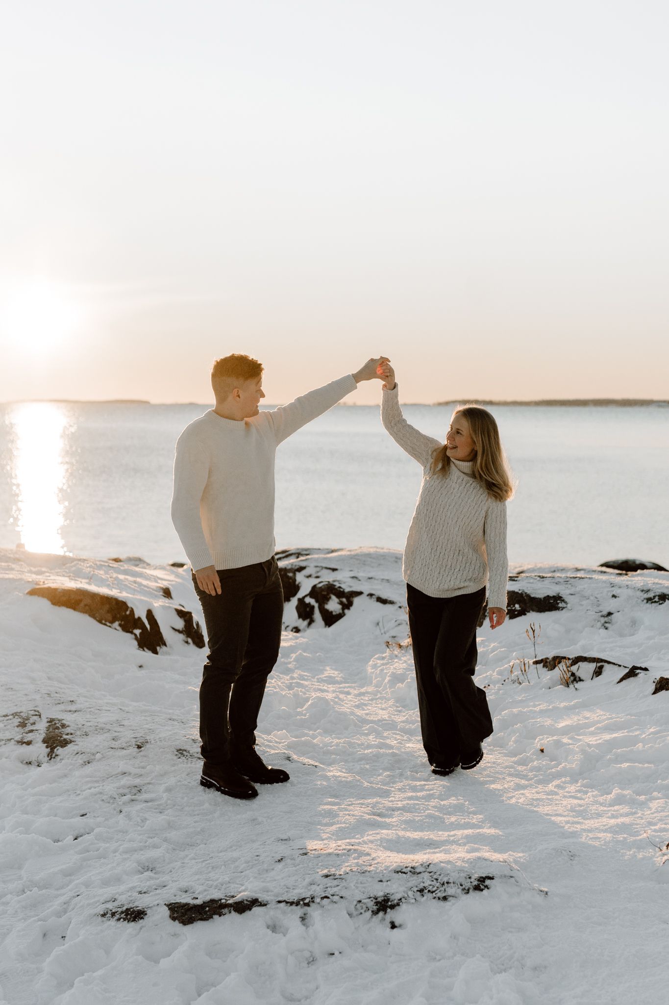 Couple in matching cream sweaters dance together on a snowy beach during a glowing winter sunset.
