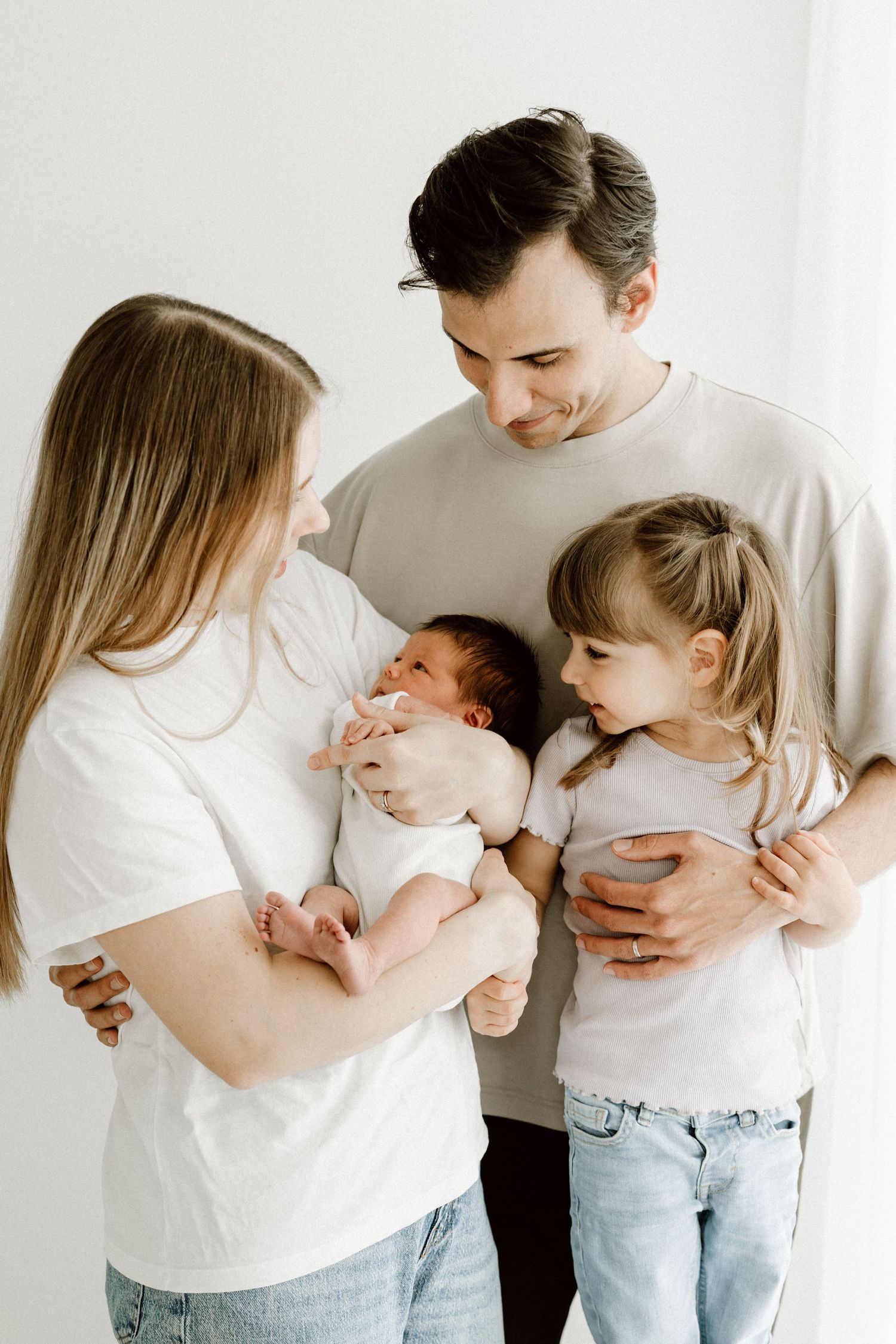 A smiling family of four in white tops stands together as mother holds their newborn baby.