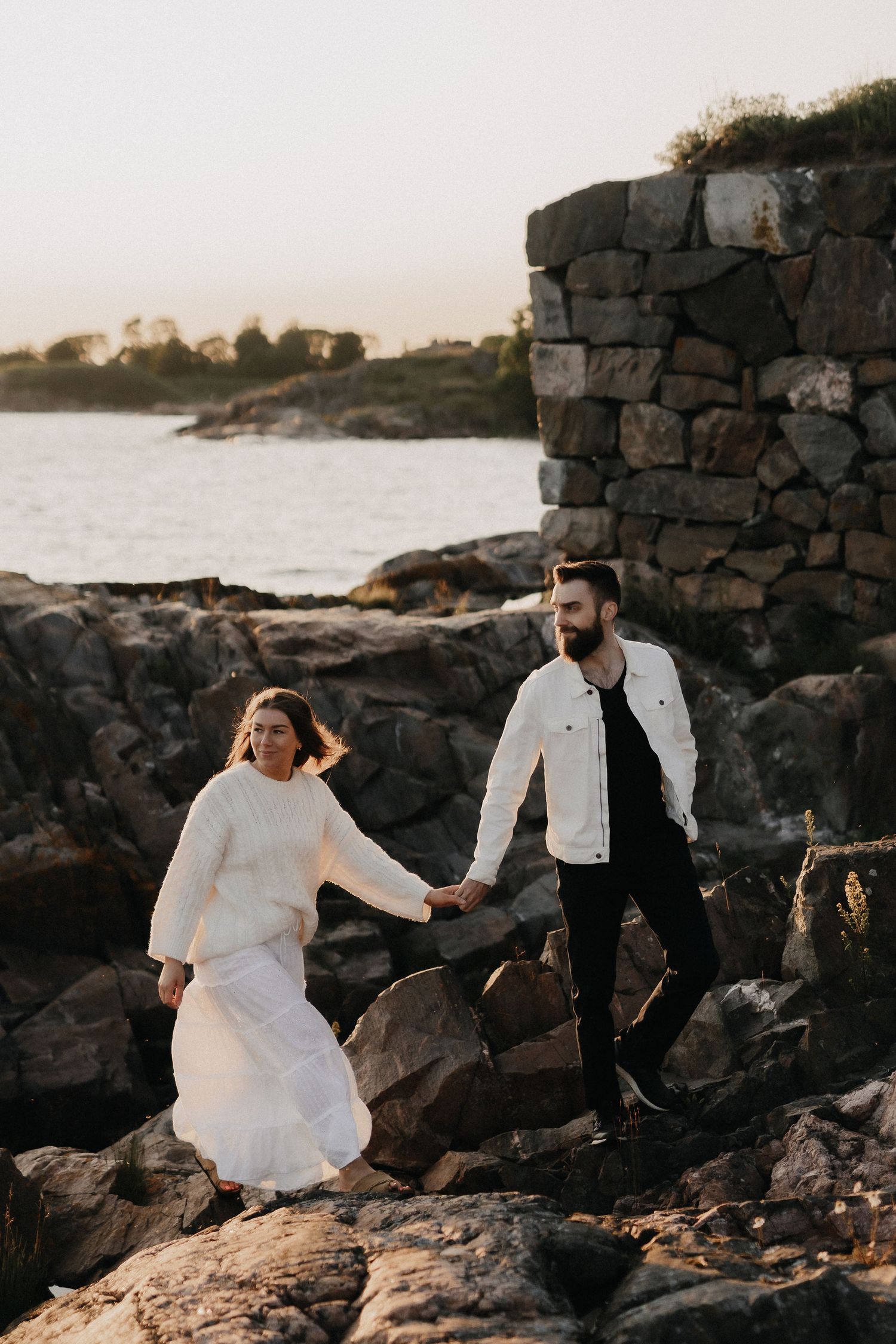 Couple holding hands while navigating rocky coastal terrain near stone ruins at golden hour.