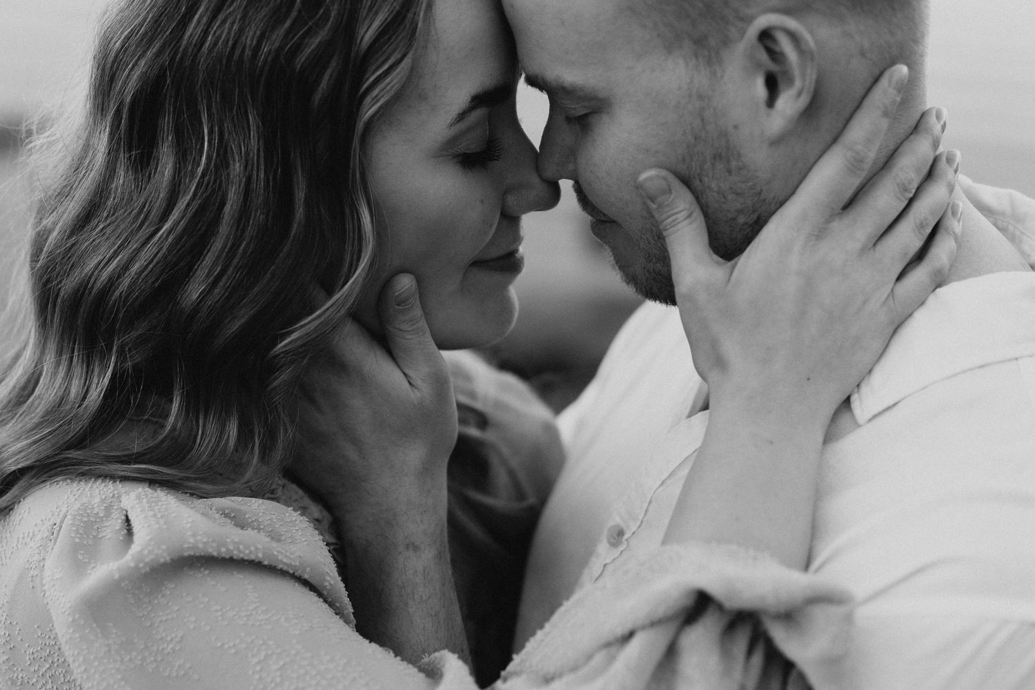 Intimate black and white close-up of couple leaning foreheads together, hands gently touching faces.