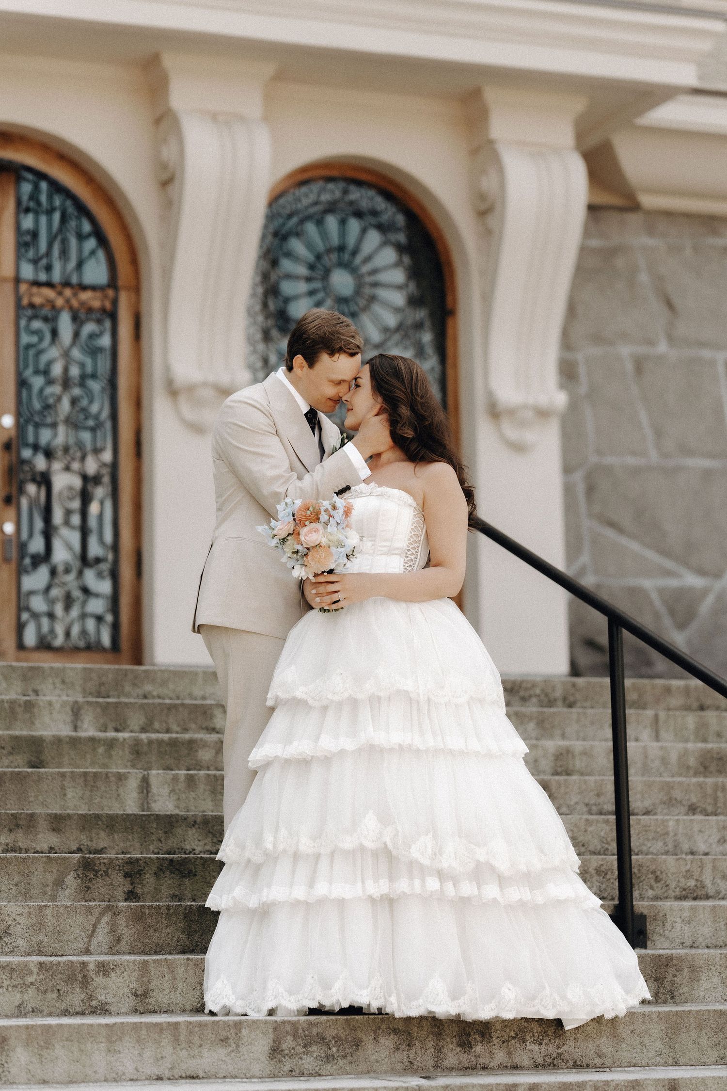 Bride and groom share romantic moment on stone steps outside elegant building with ornate doors.