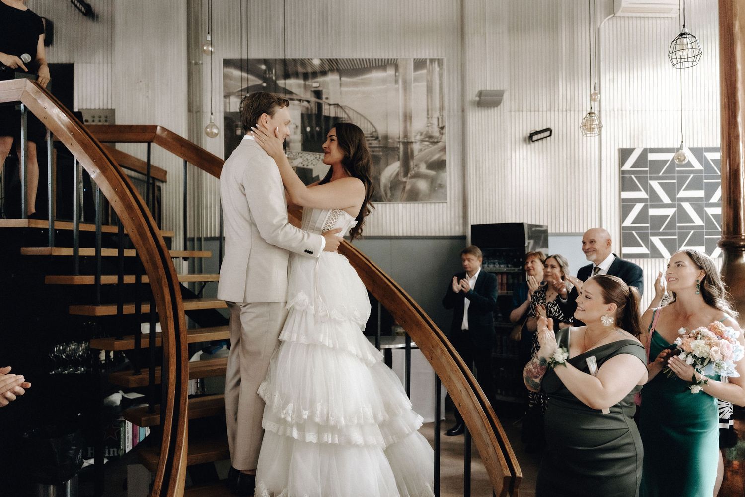 Bride and groom share a romantic embrace on a spiral staircase while wedding guests applaud below.