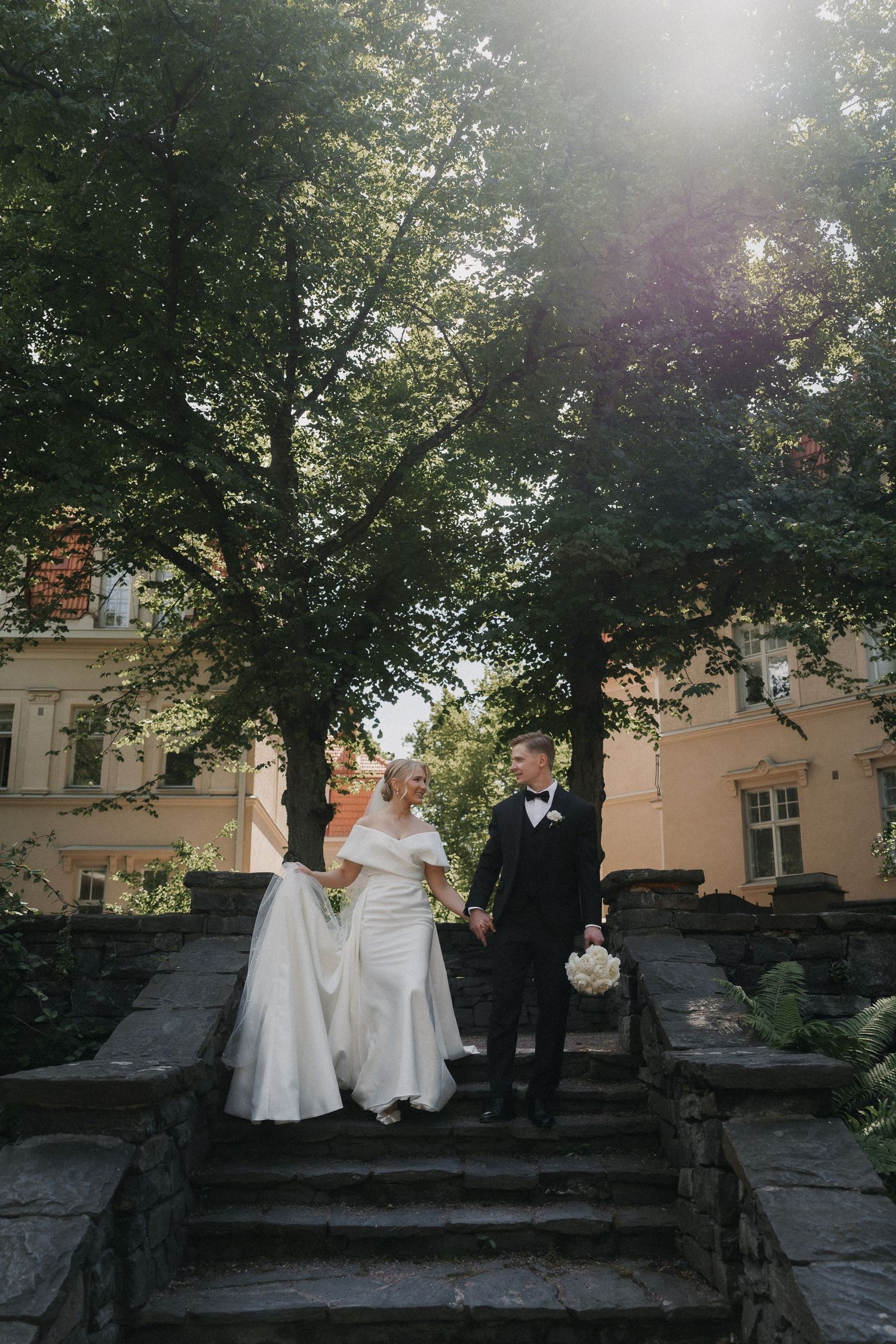 Bride and groom walk down stone steps hand in hand beneath sunlit trees near a historic building.