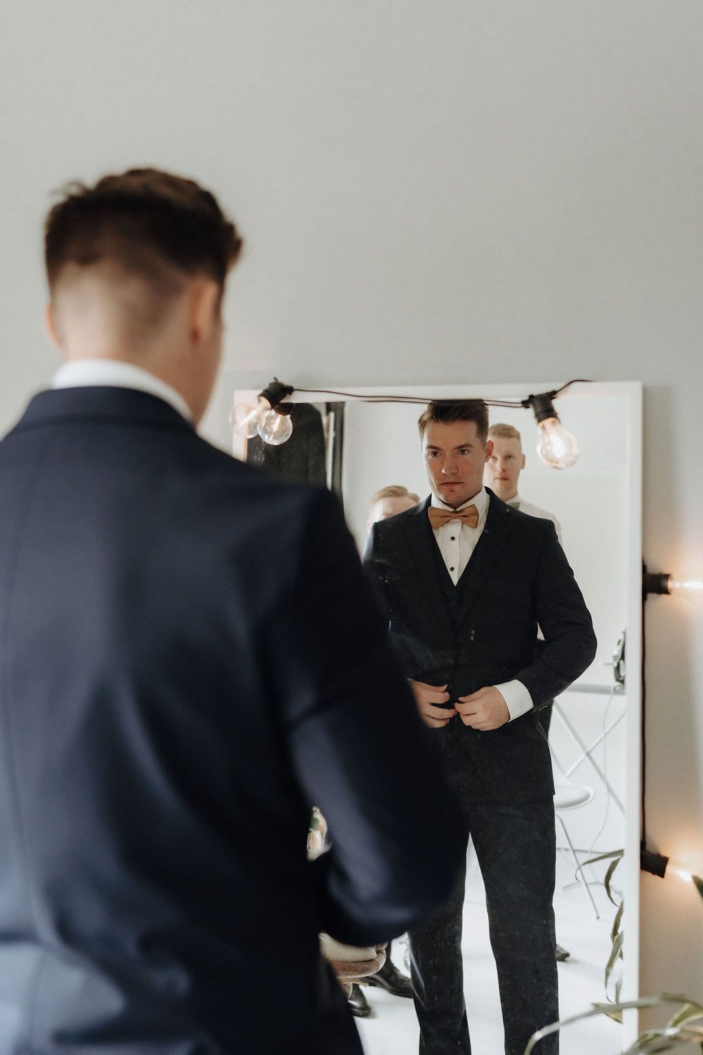 Dapper groom in navy tuxedo with bow tie checking appearance in mirror.