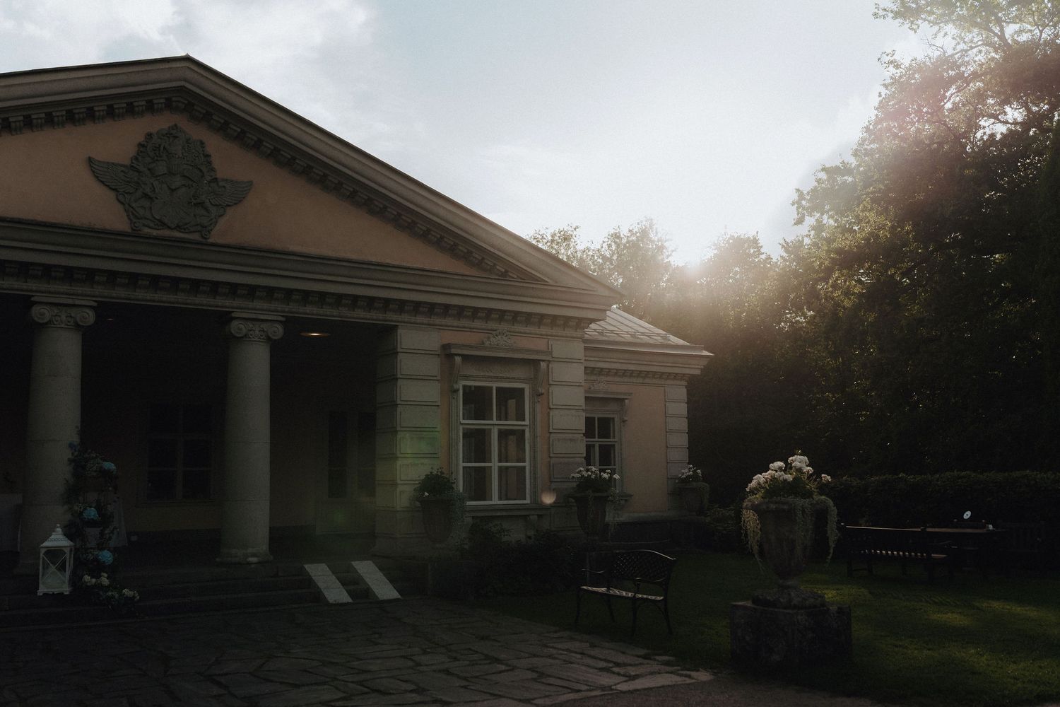 Sunlit neoclassical wedding venue exterior with columns surrounded by lush green trees.
