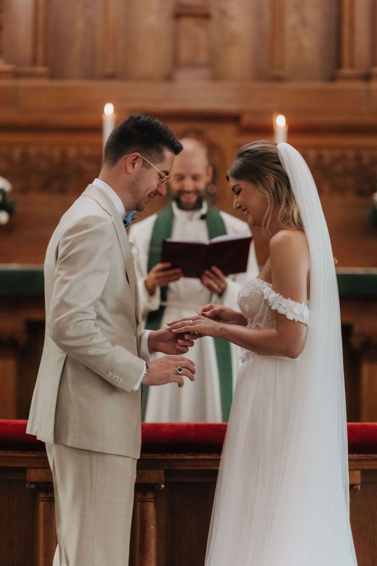 Groom placing wedding ring on brides finger as smiling officiant reads from book at candlelit church altar.