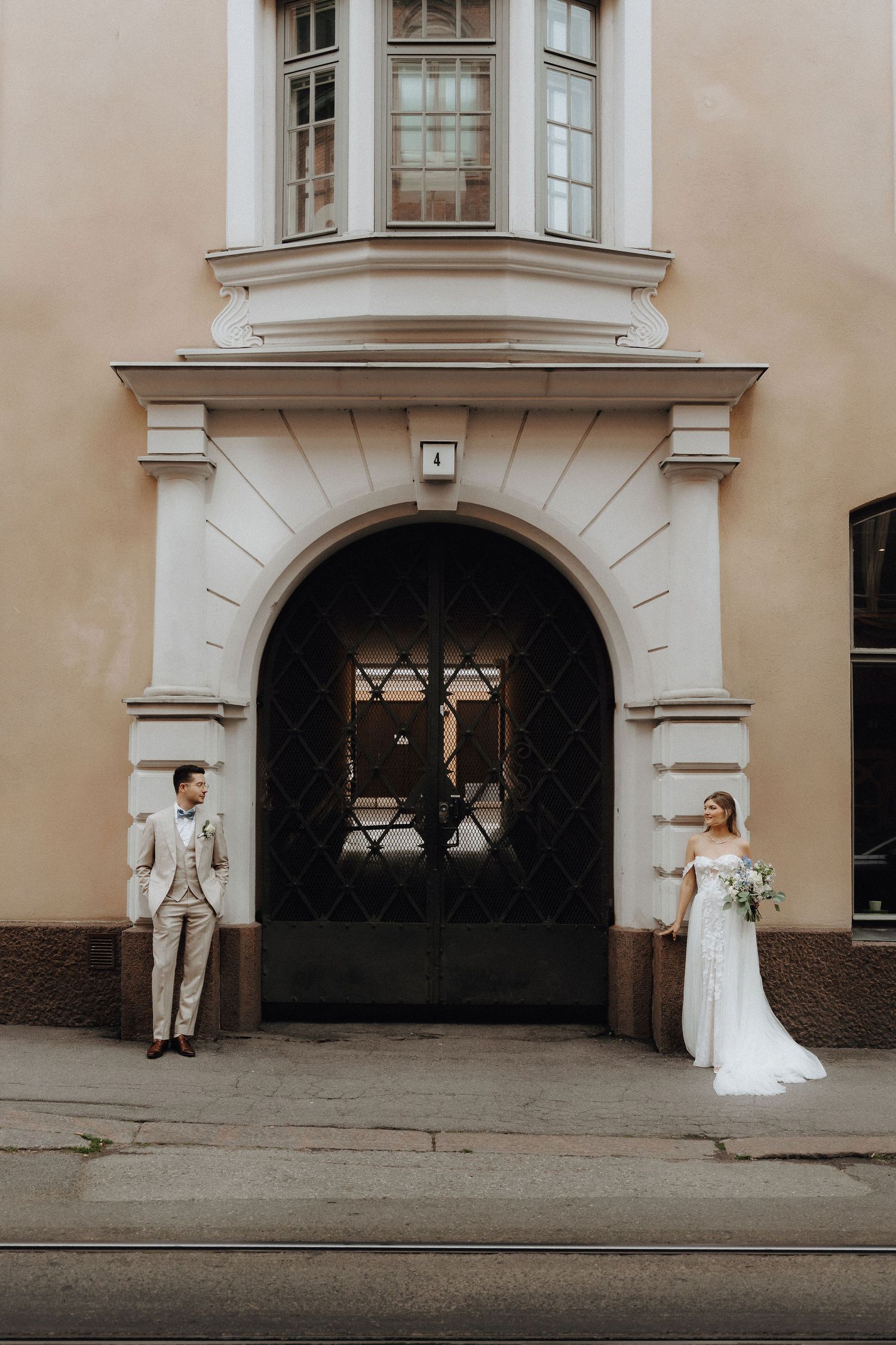 Bride in white gown and groom in beige suit posing on opposite sides of an arched black door.