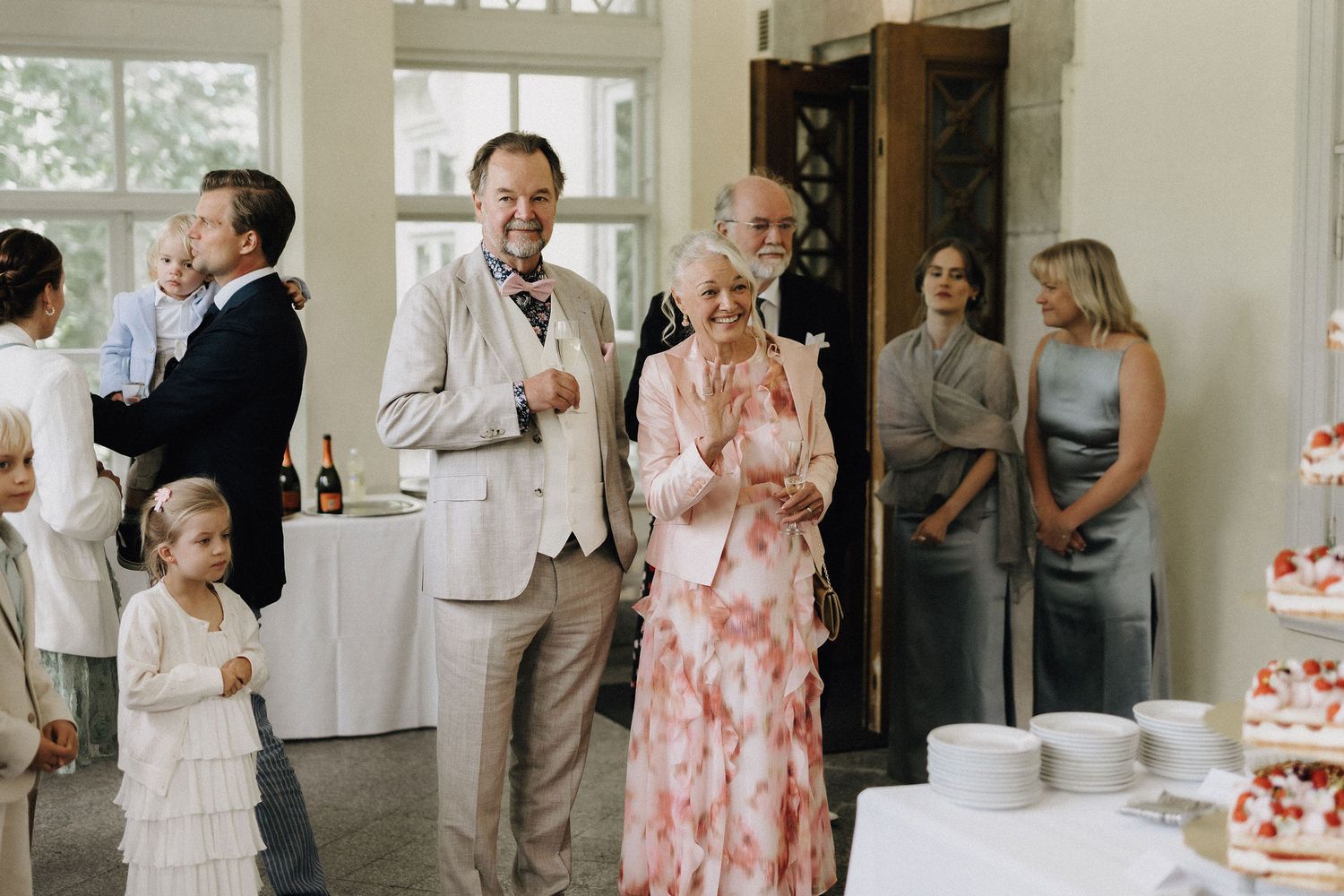 Joyful wedding guests including an older couple and children gathered around a cake table indoors.