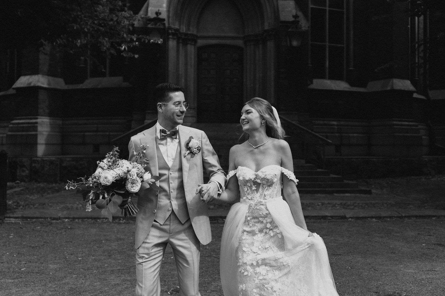 Laughing bride and groom in elegant attire walking together outside a historic building in black and white.