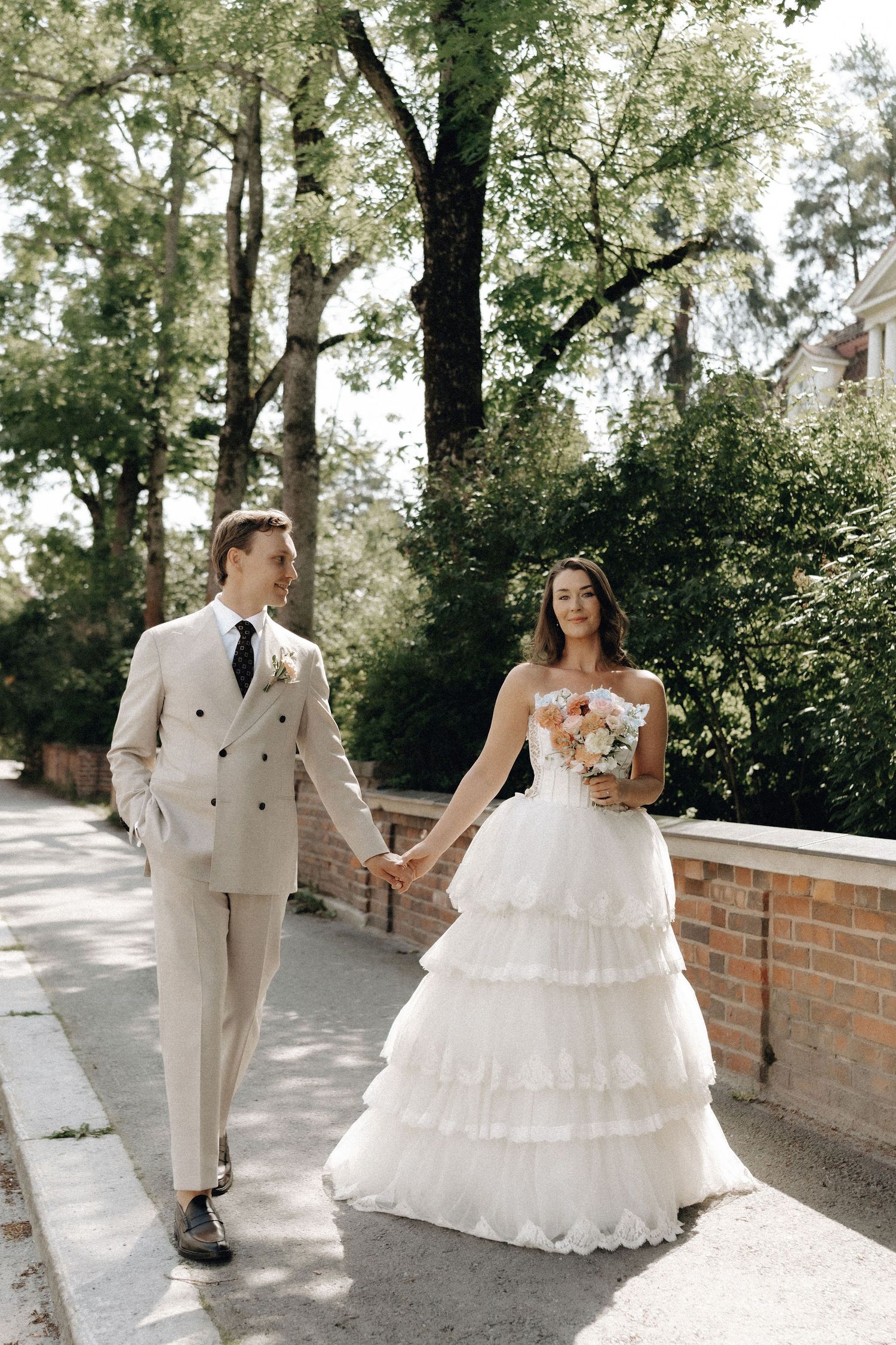 Bride in layered white gown and groom in beige suit stroll together along a brick-lined garden path.