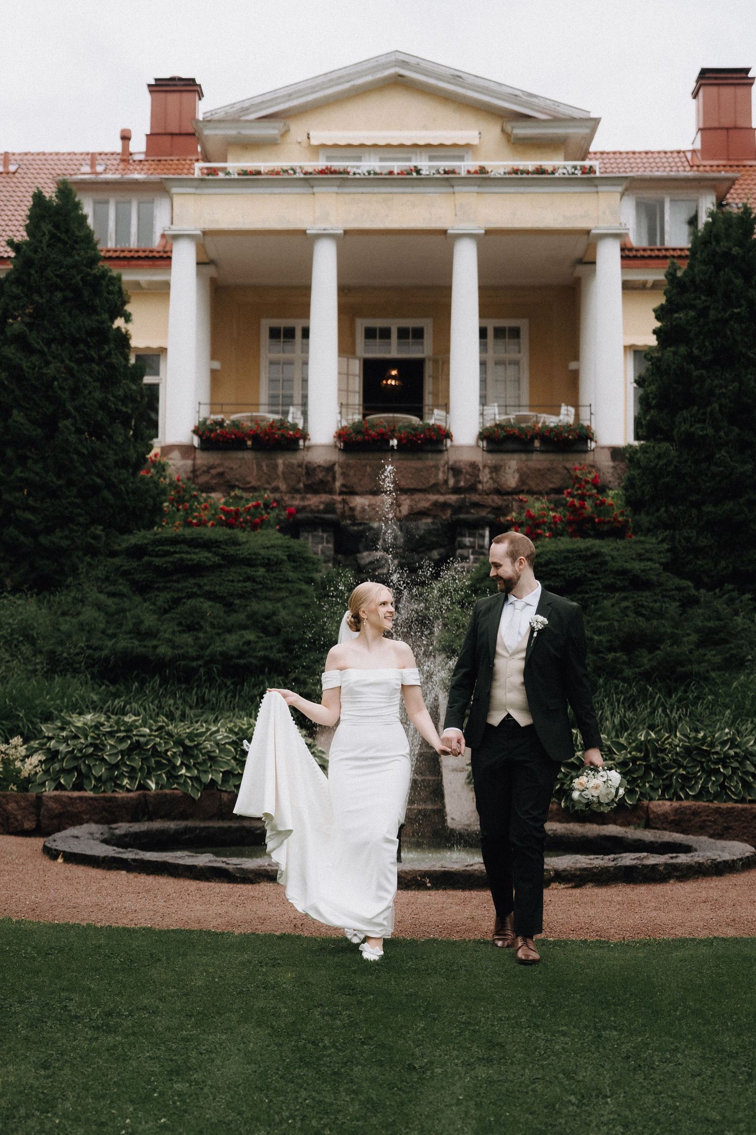 Bride and groom walking hand in hand in front of elegant estate fountain.