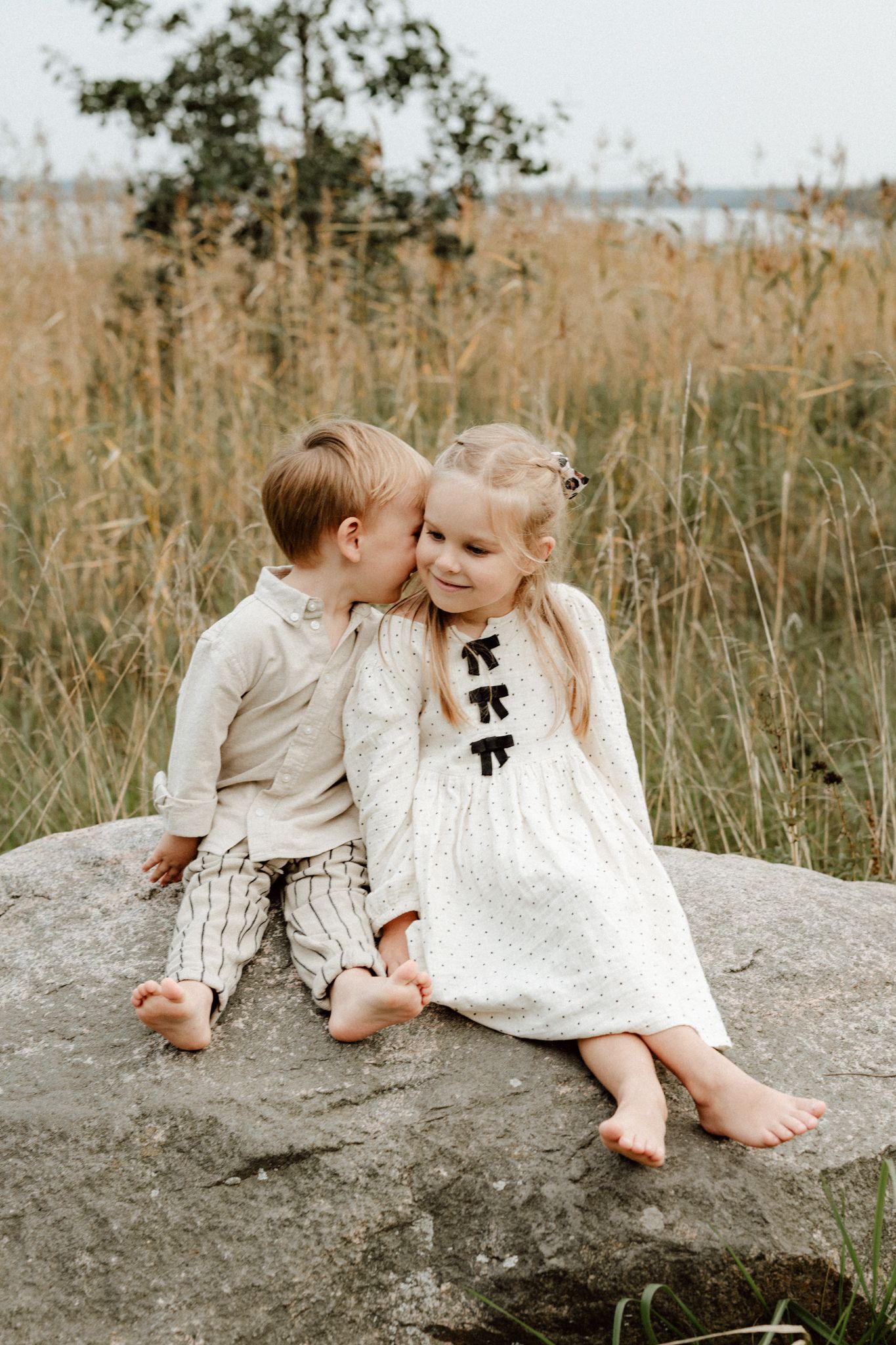 Young boy whispering to blonde girl sitting together on rock in golden grass field.