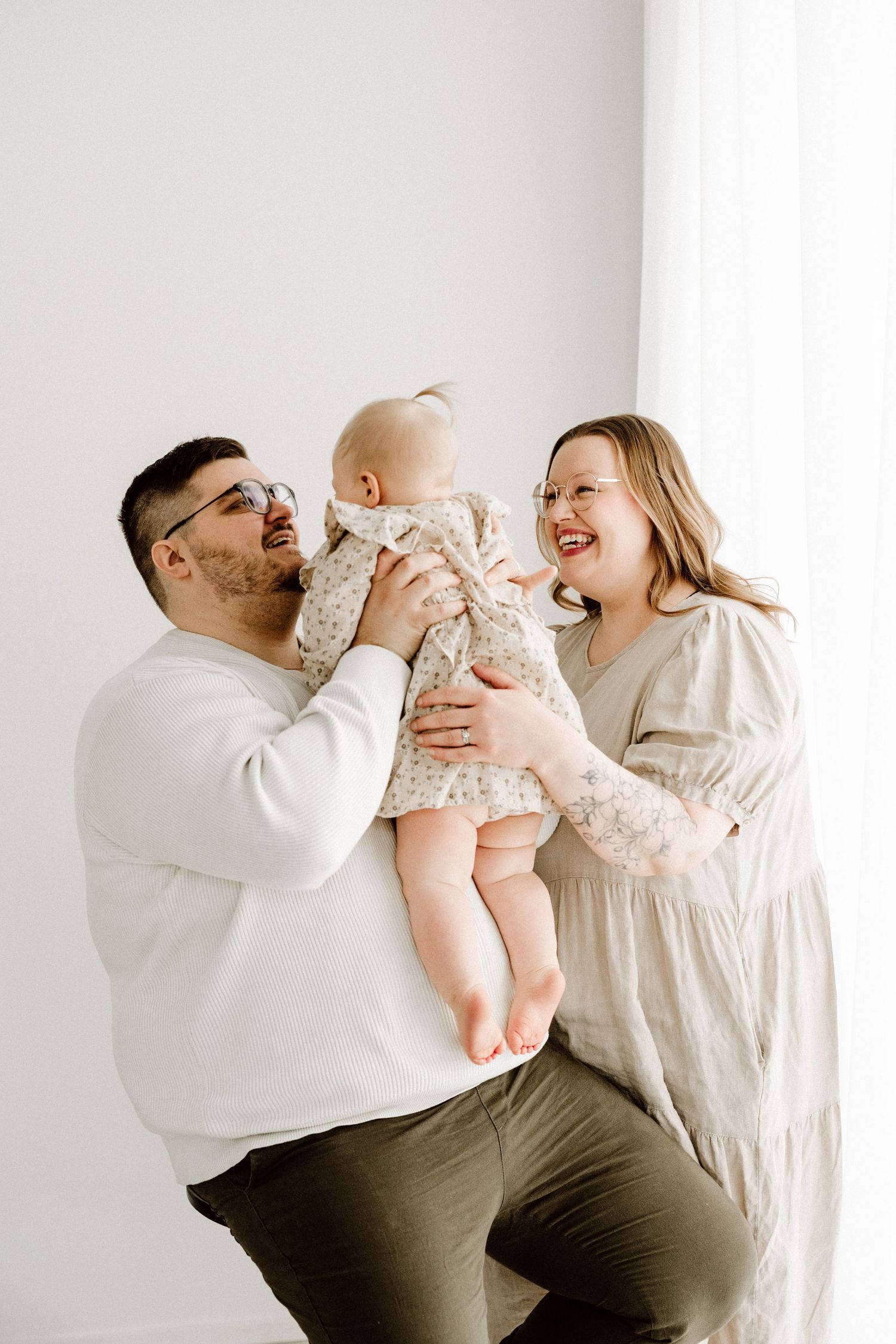 Two parents in neutral tones smile while holding their baby girl near a bright window in a studio.