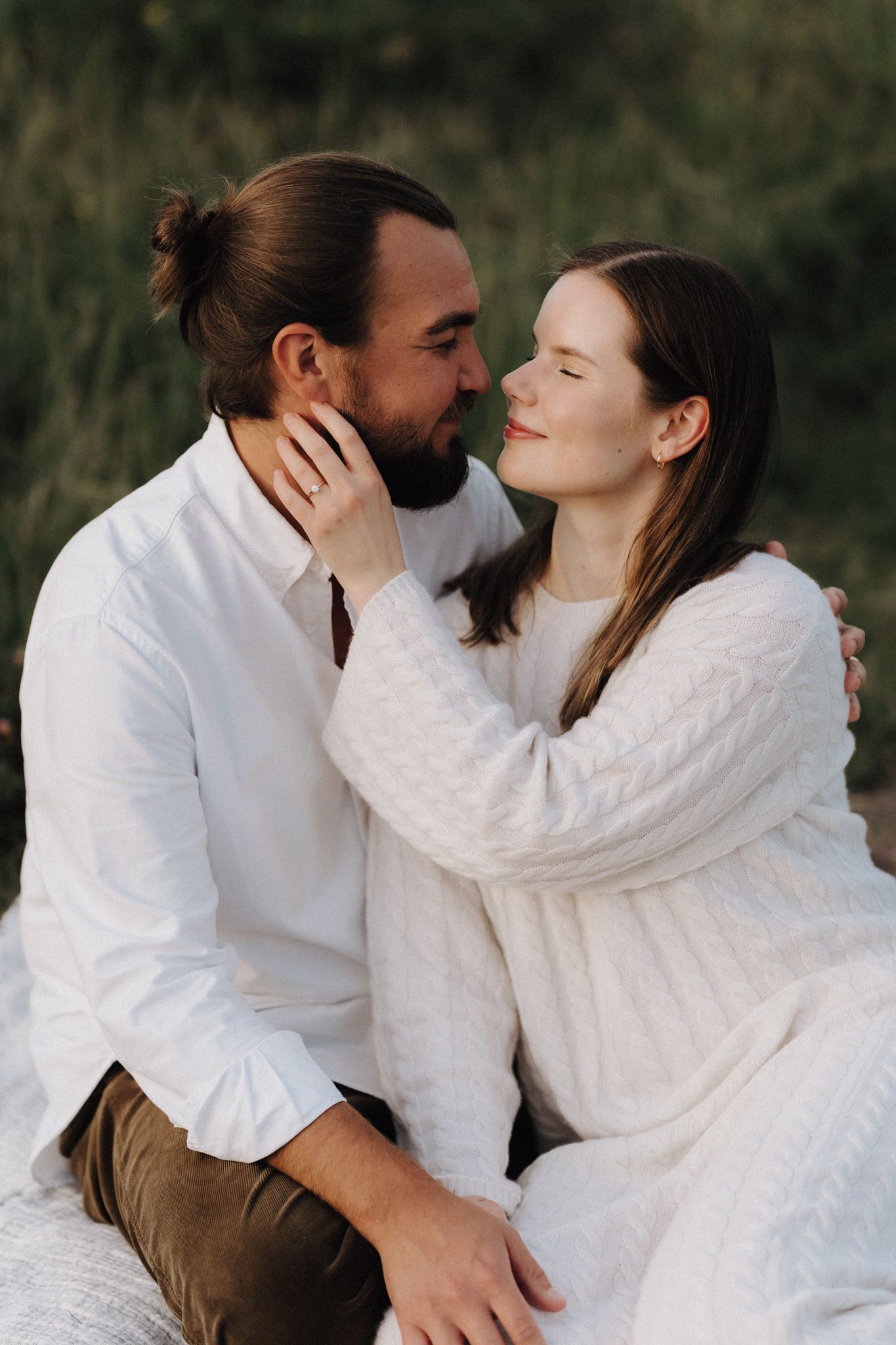 A couple sits closely together outdoors, about to share a gentle kiss at sunset.