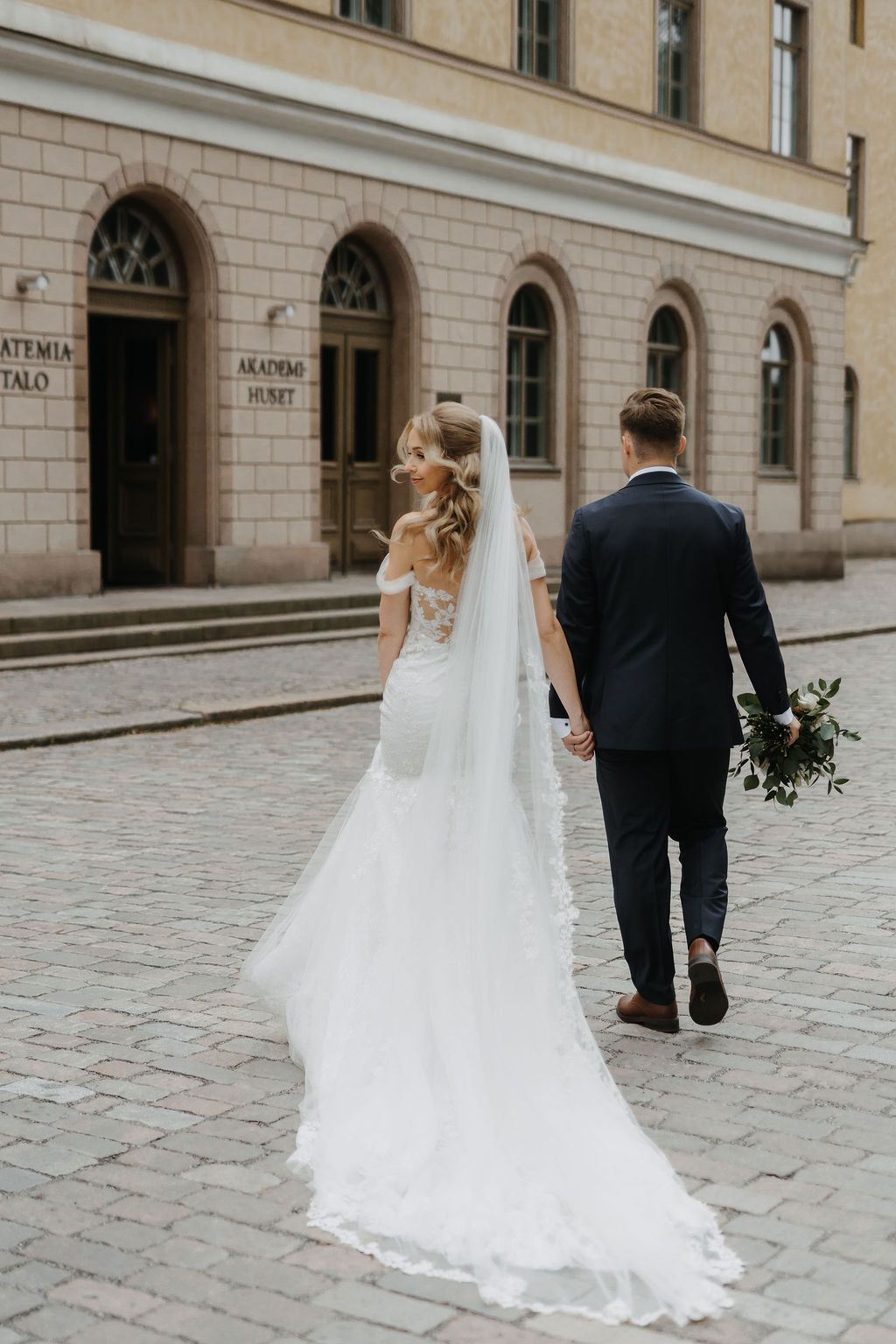 Bride in flowing lace wedding gown and long veil walks with groom past Akademia Talo on cobblestones.