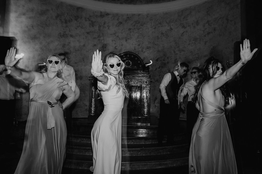 Black and white photo of bride and bridesmaids in sunglasses dancing with hands raised at wedding.
