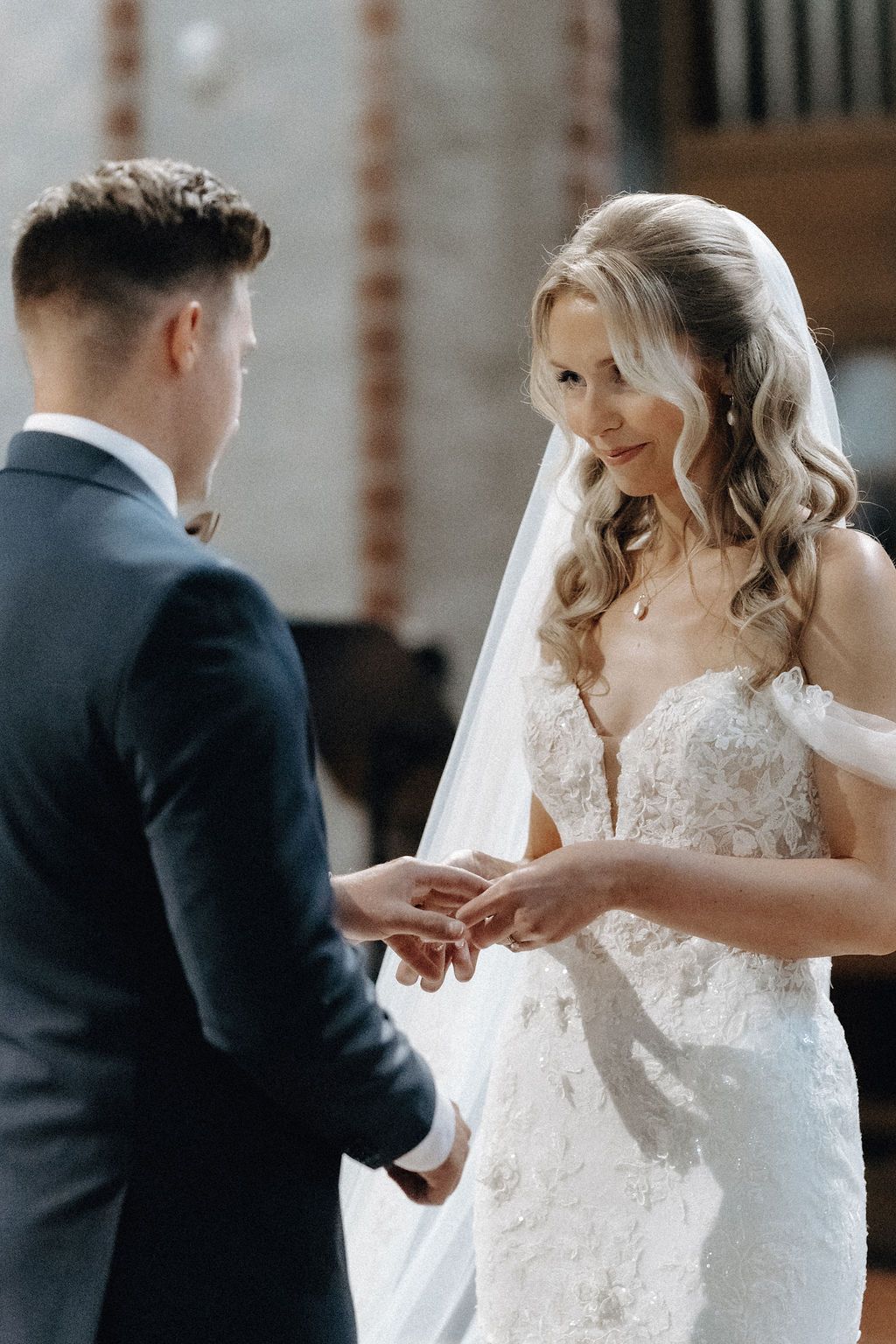 Bride in lace wedding gown places ring on grooms finger during church ceremony.