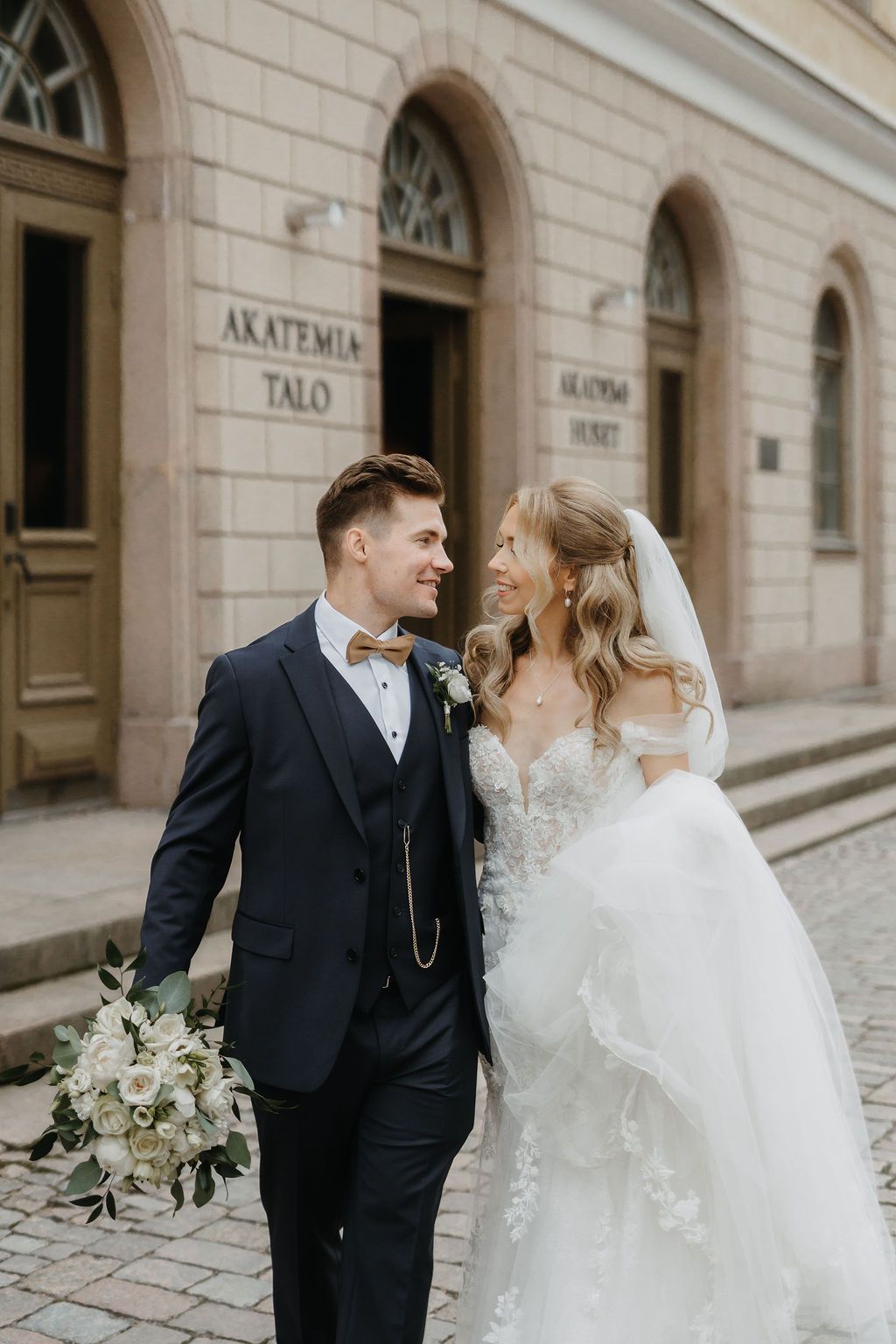 Bride and groom gazing at each other lovingly outside Akatemia-Talo, groom holding white floral bouquet.