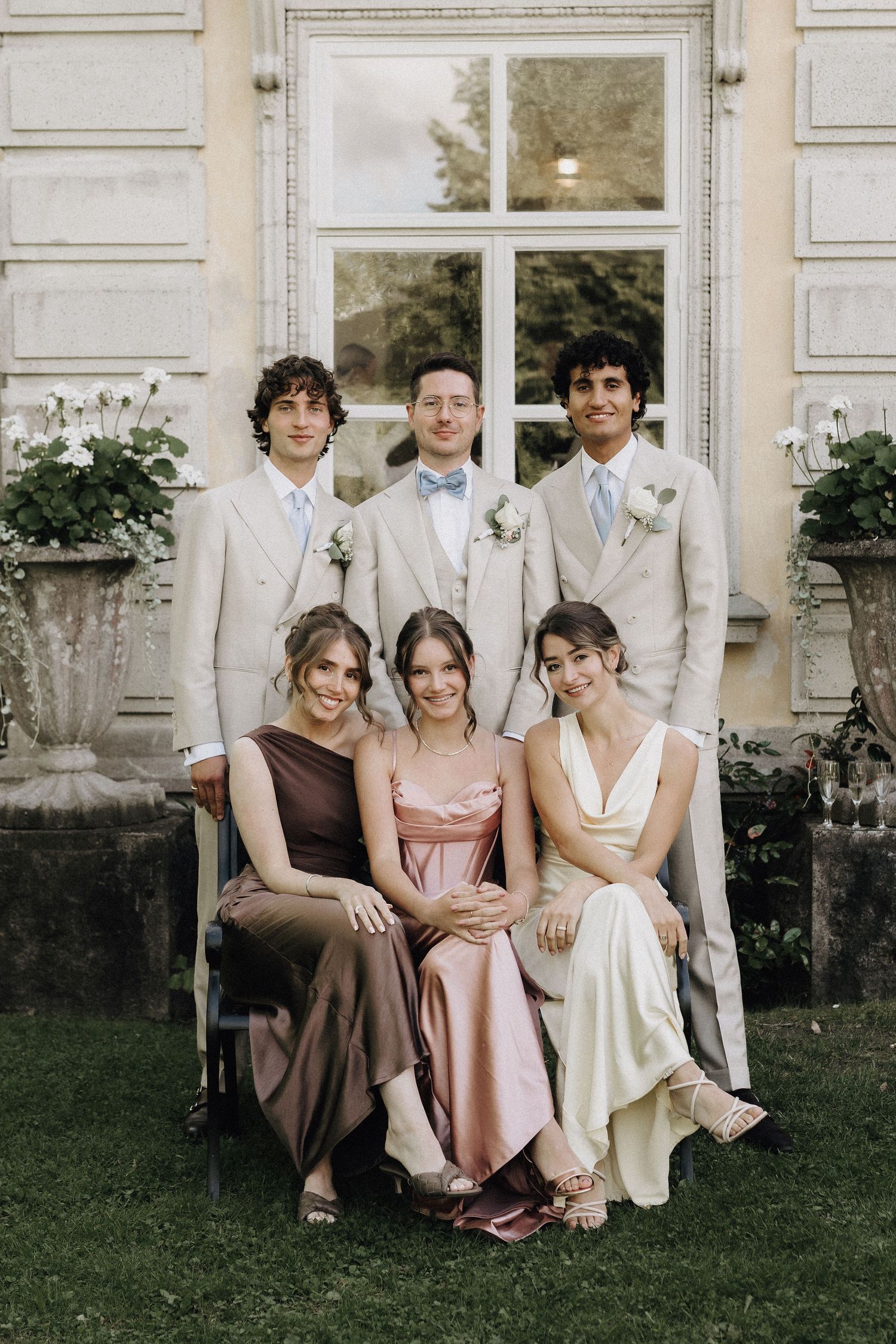 Three groomsmen in beige suits with three bridesmaids in satin dresses posing for wedding photo.