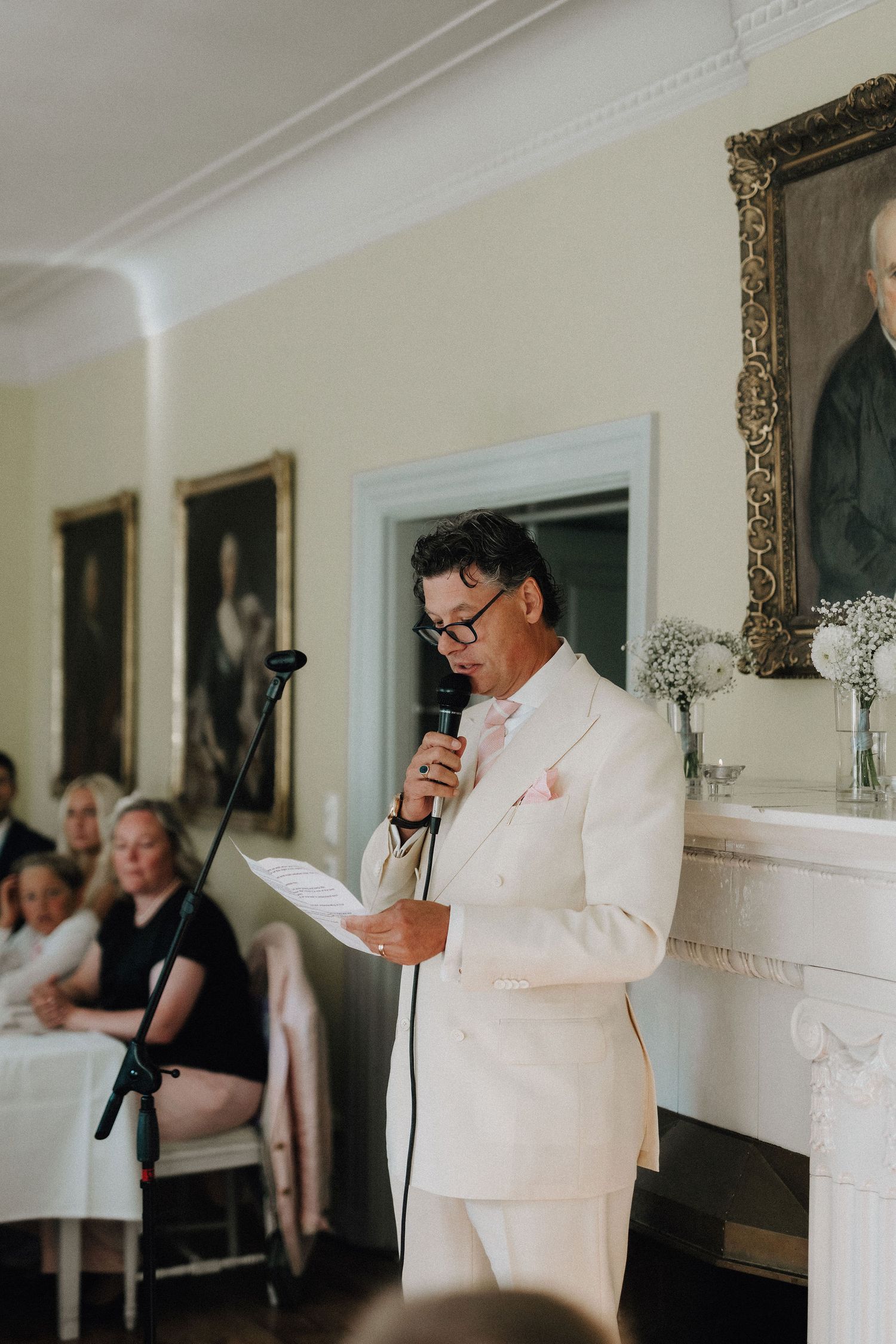 Elegant man in white suit delivers speech at wedding reception in a classical interior with portrait paintings.